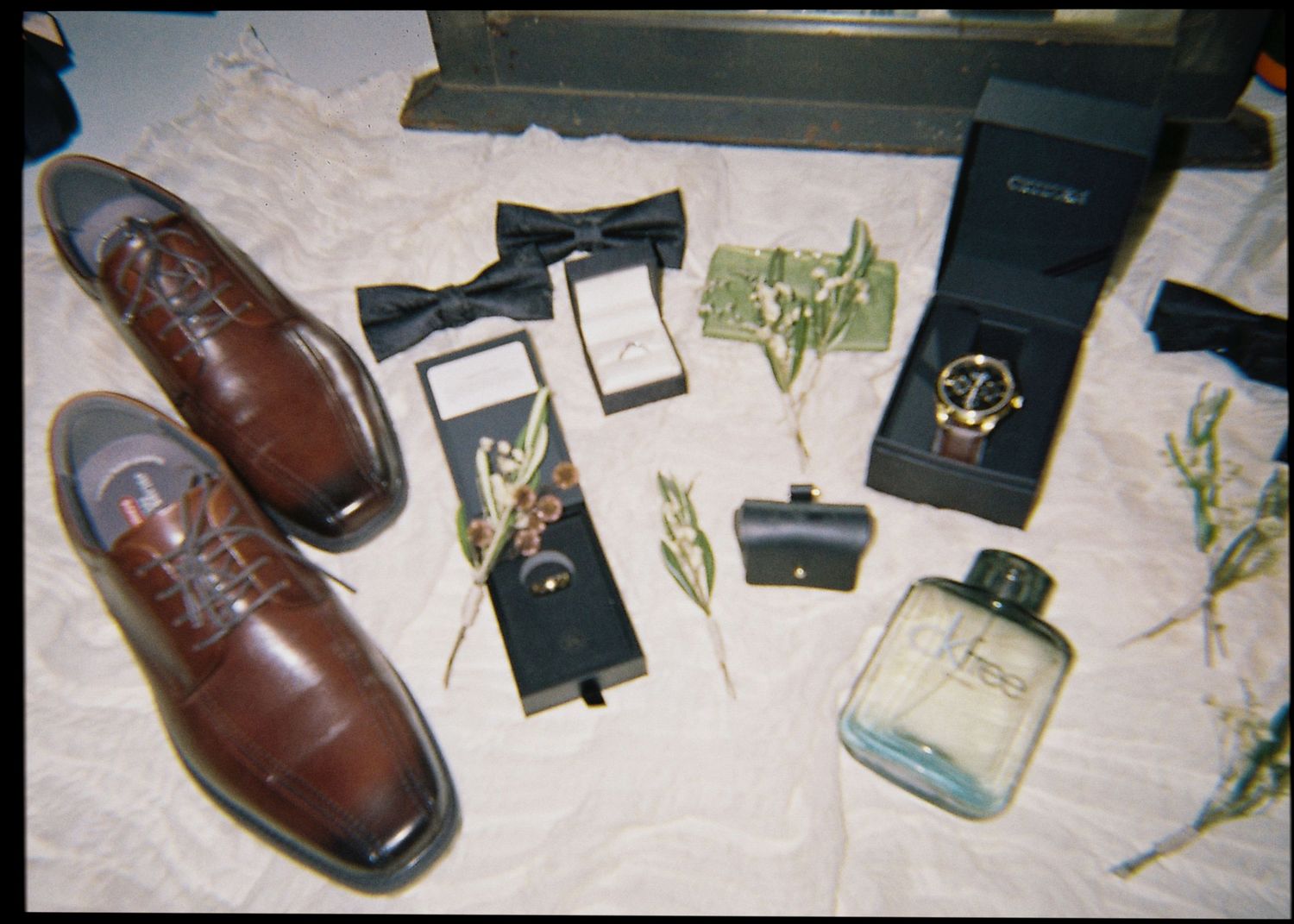 Groom wedding accessories flatlay featuring brown leather shoes, bow tie, watch, and cologne.