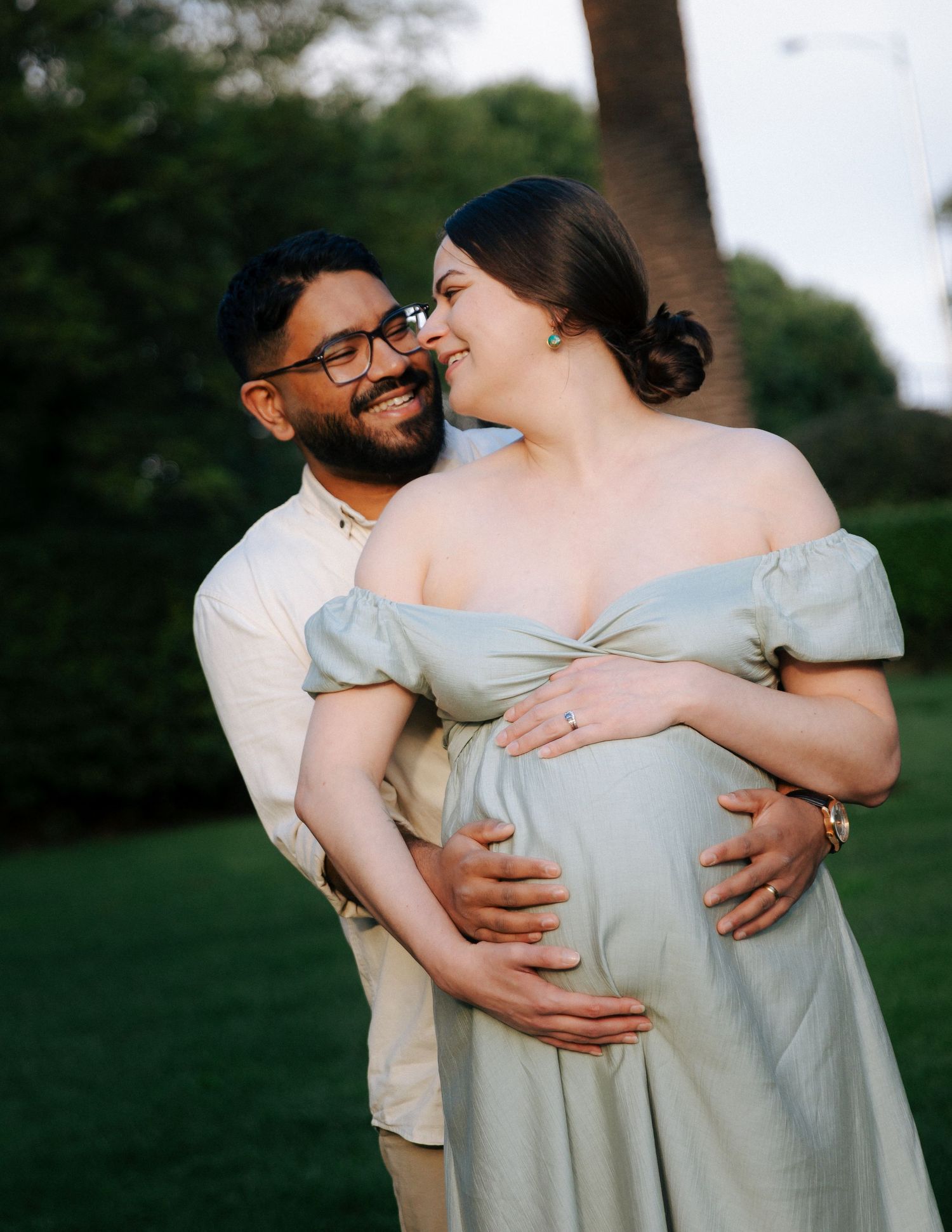 Smiling couple sharing a joyful maternity photo in a lush park, man embracing pregnant woman in sage dress.