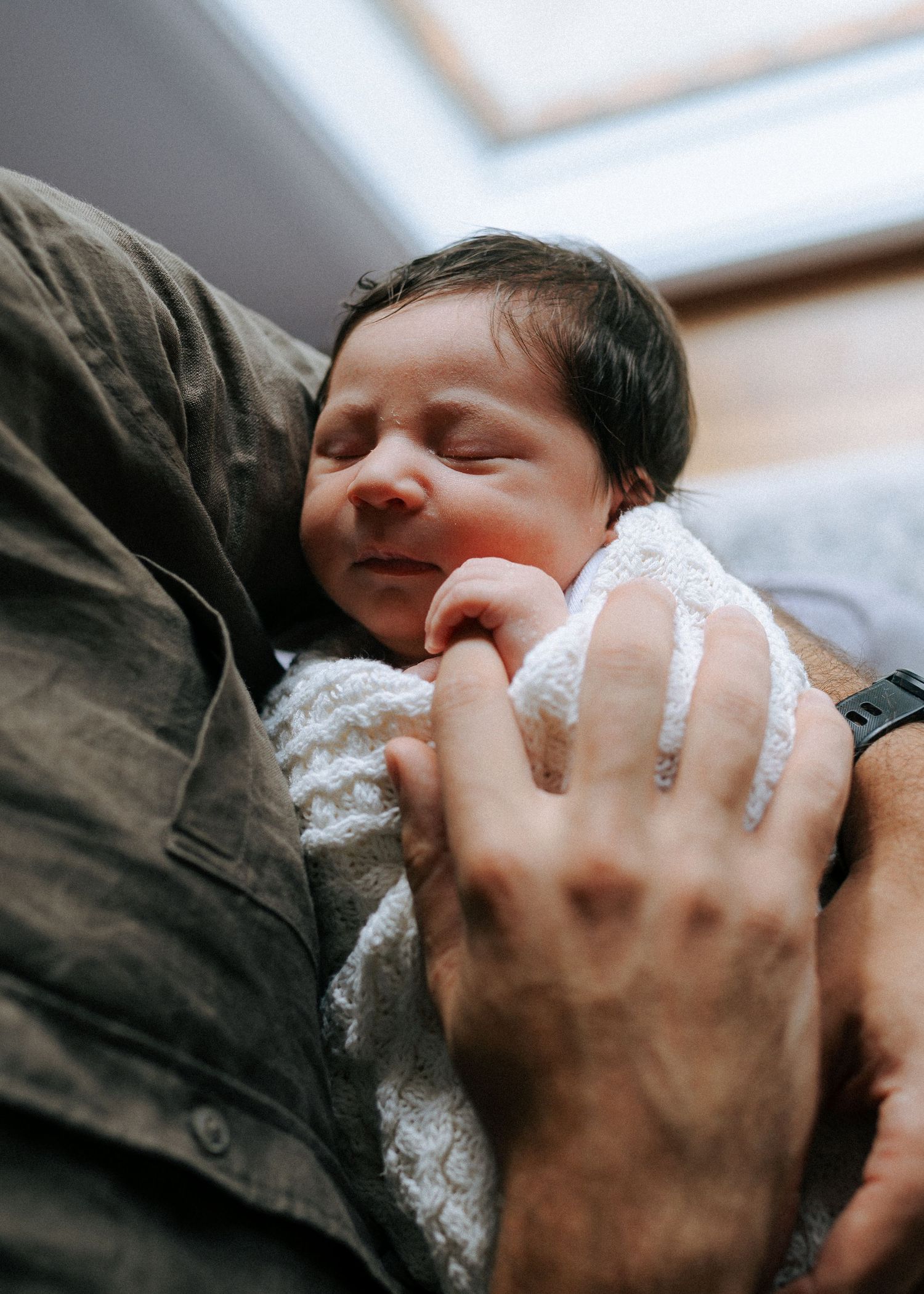 Close-up of sleeping dark-haired newborn baby wrapped in white lace blanket in fathers arms.