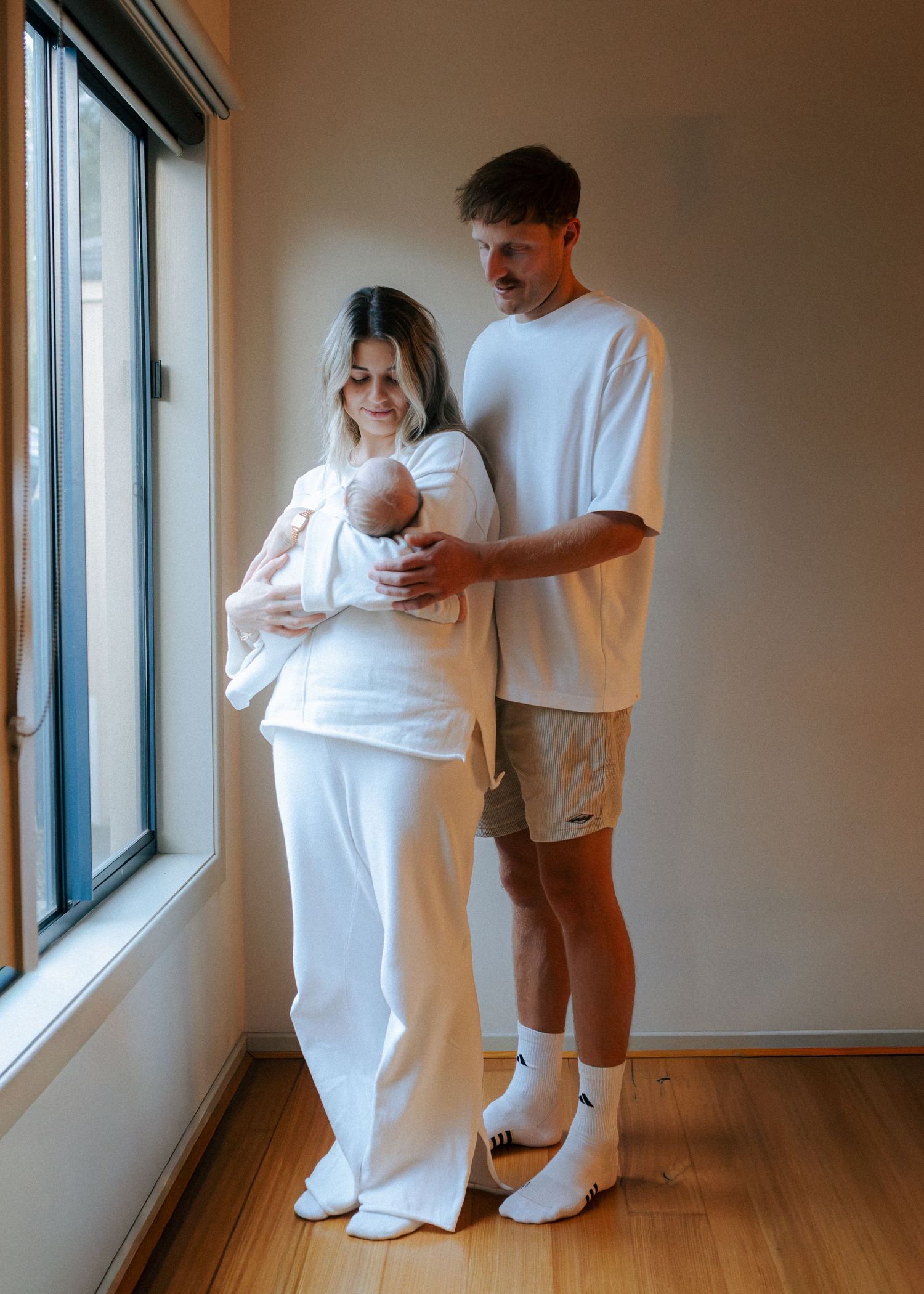 New parents in white outfits standing by window lovingly looking at their newborn baby.