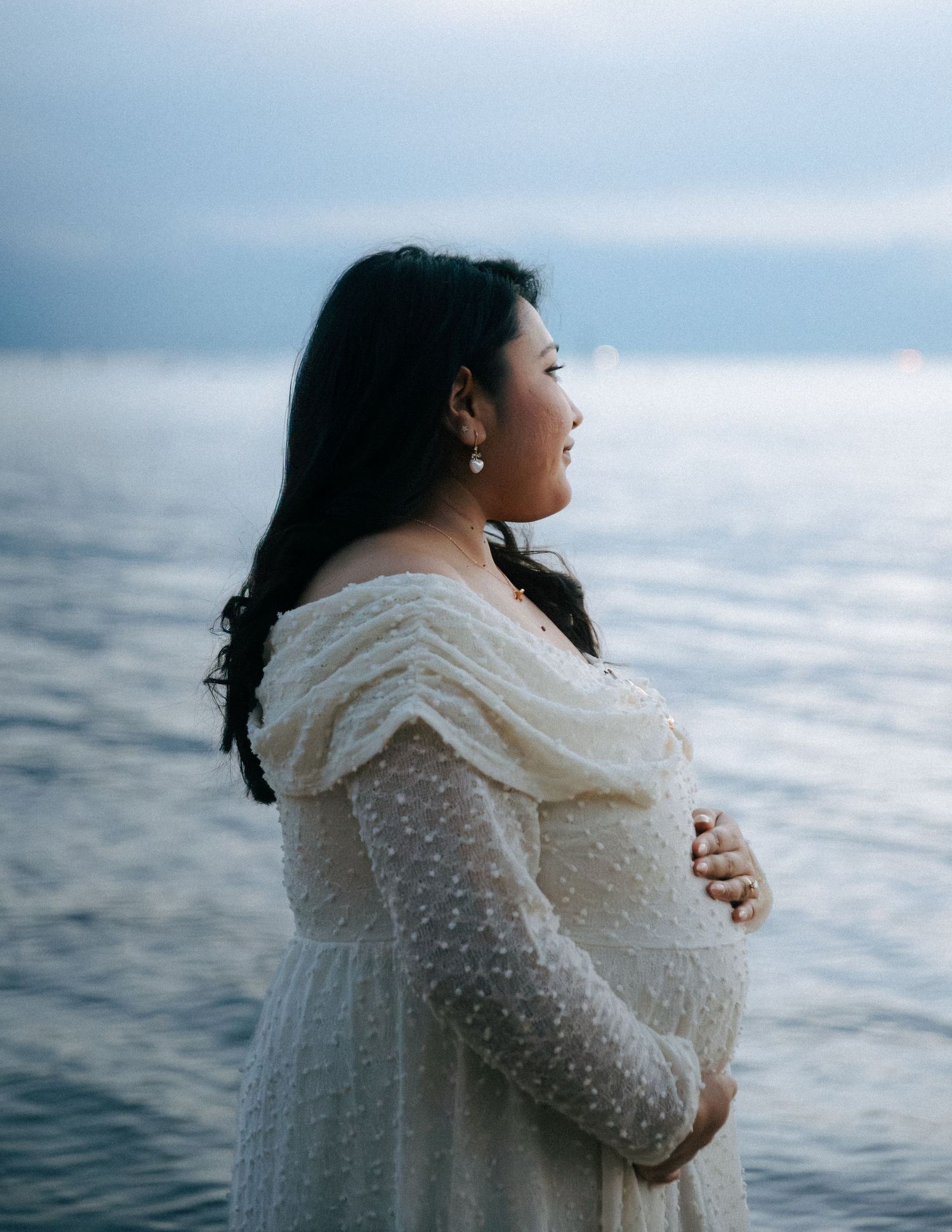 Pregnant woman in white off-shoulder dress gazing at ocean, cradling baby bump at dusk.