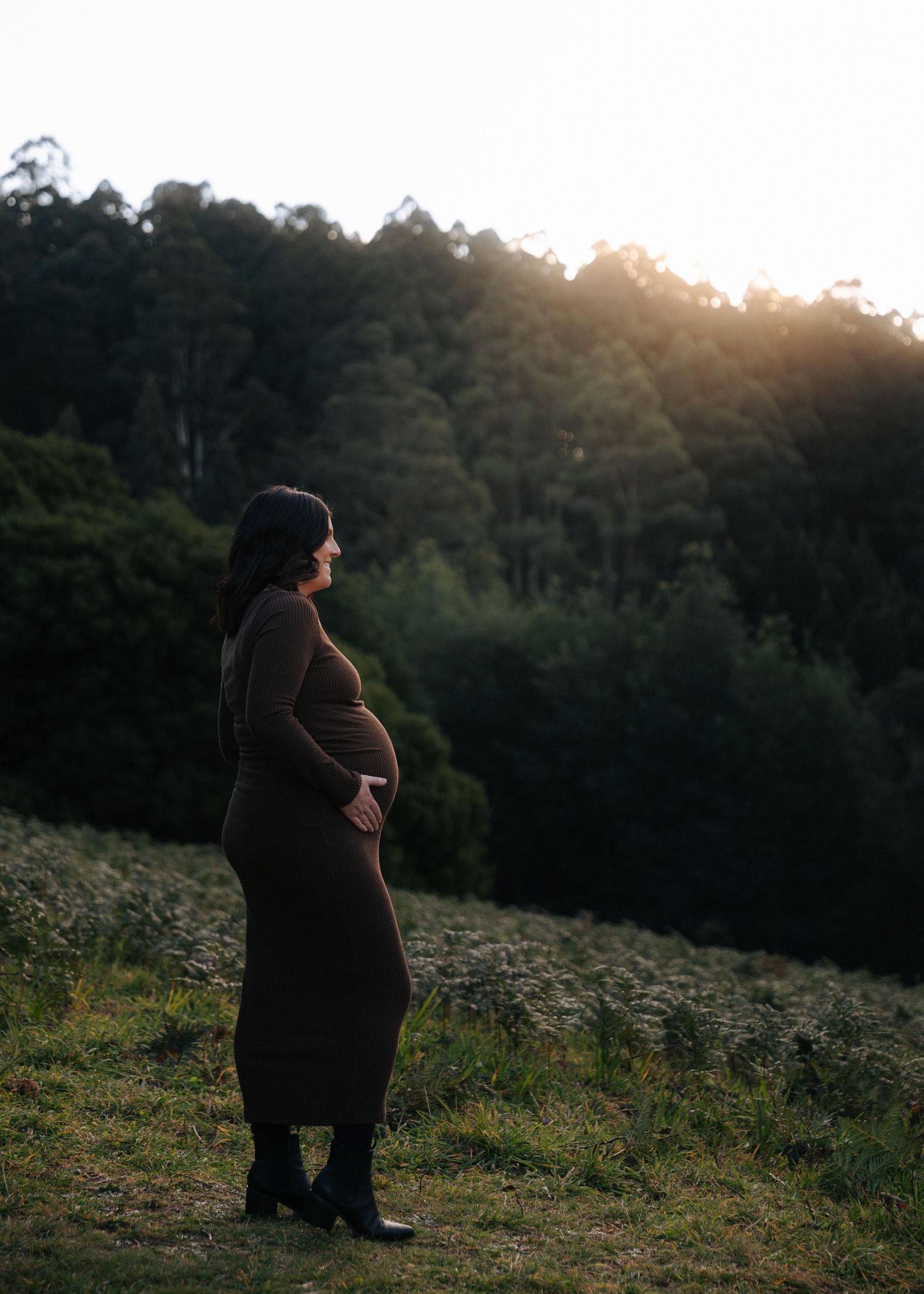 Pregnant woman in brown maxi dress standing alone in a field gazing into the distance at dusk.