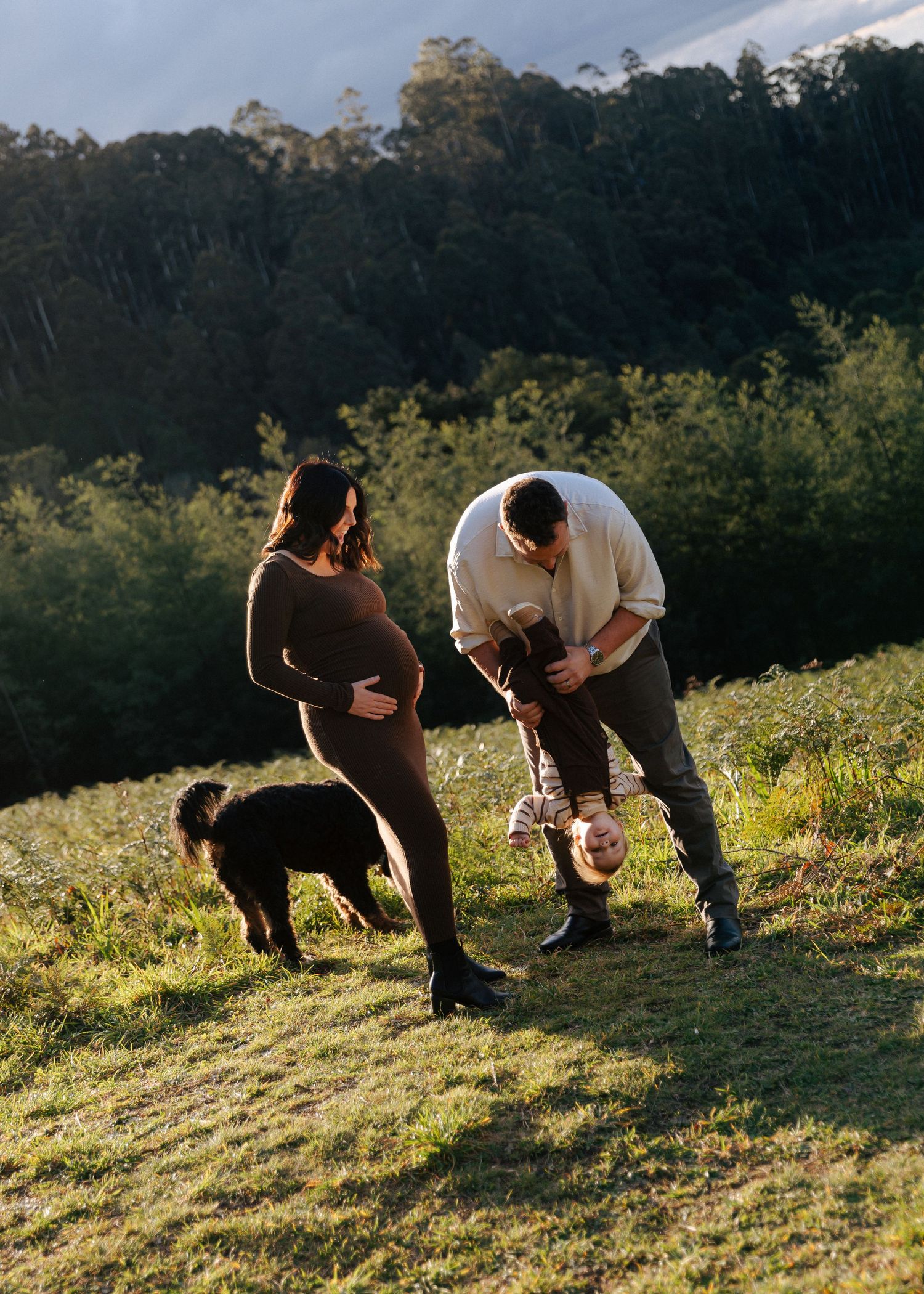 Pregnant woman watches as father playfully holds toddler upside down on sunny green hillside.