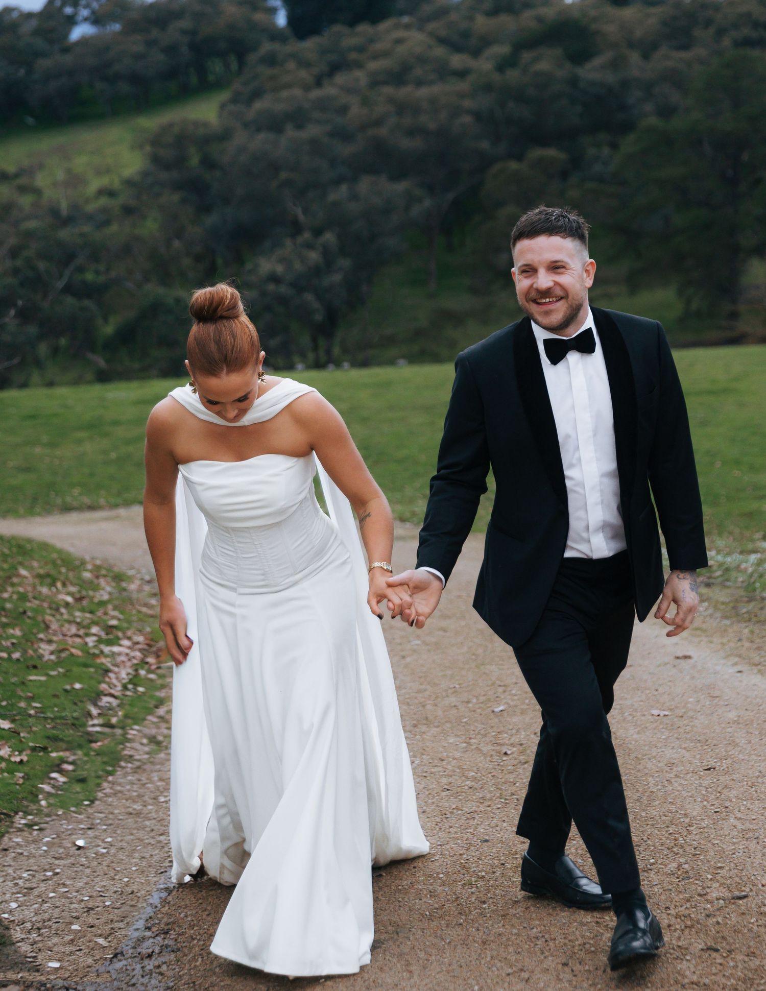 Smiling groom in black tuxedo and bride in white gown walking together on outdoor pathway.