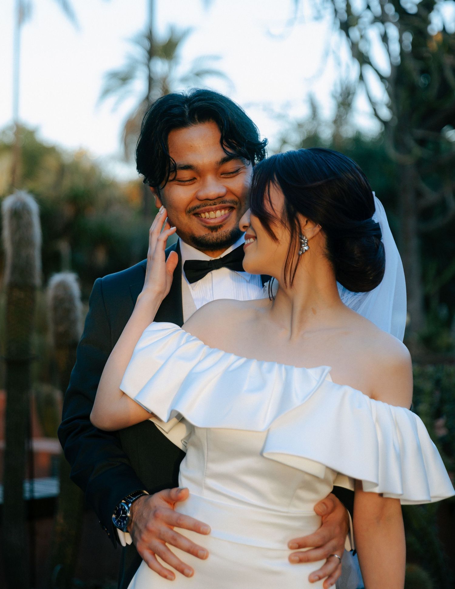 Joyful groom in tuxedo smiling as bride in off-shoulder white dress tenderly touches his face outdoors.