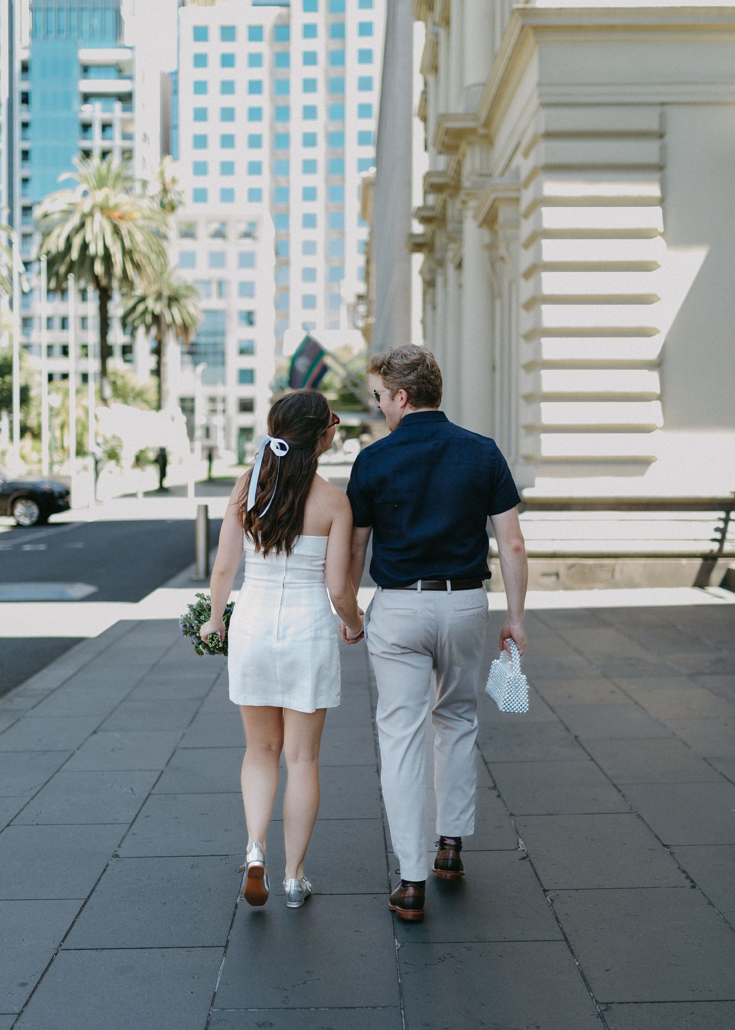 Newlywed couple walking hand-in-hand down a sunny city sidewalk, bride in white mini dress holding bouquet.