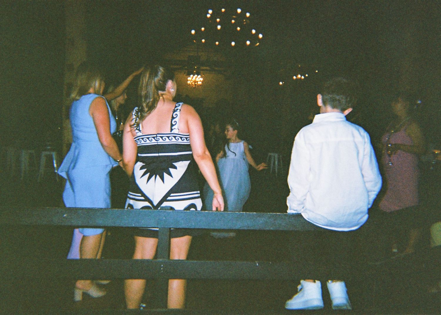 Wedding reception guests dancing together in a dimly lit rustic barn venue with chandeliers.