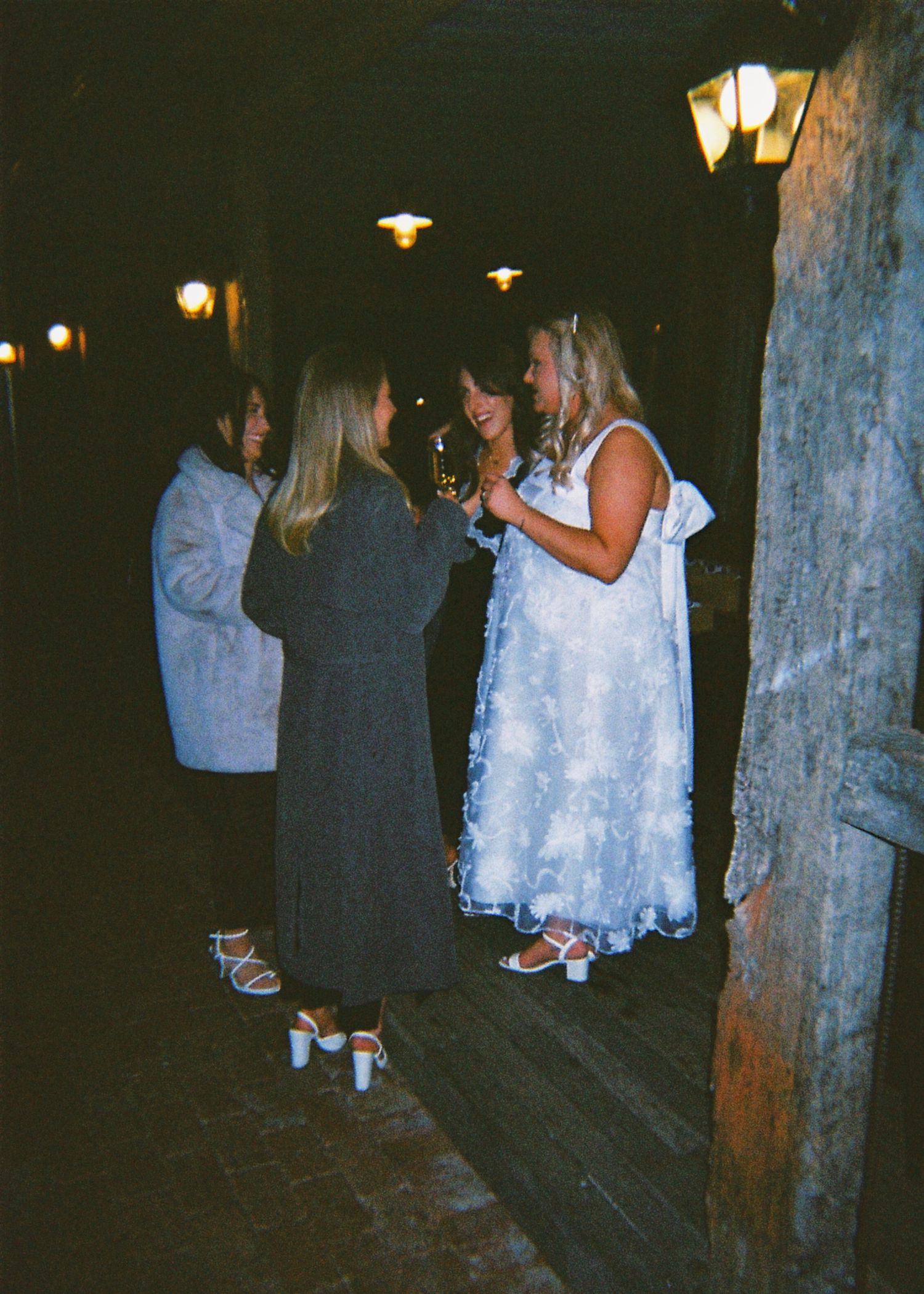 Bride in white floral gown chatting with guests under outdoor lights at a nighttime wedding reception.