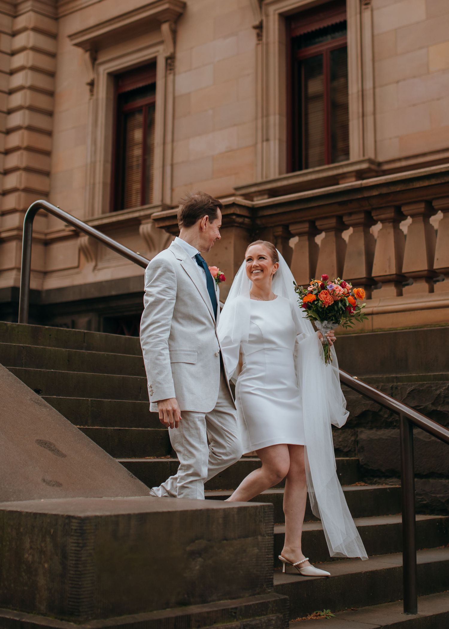Smiling bride in short white dress and groom in tan suit descend sandstone building steps.