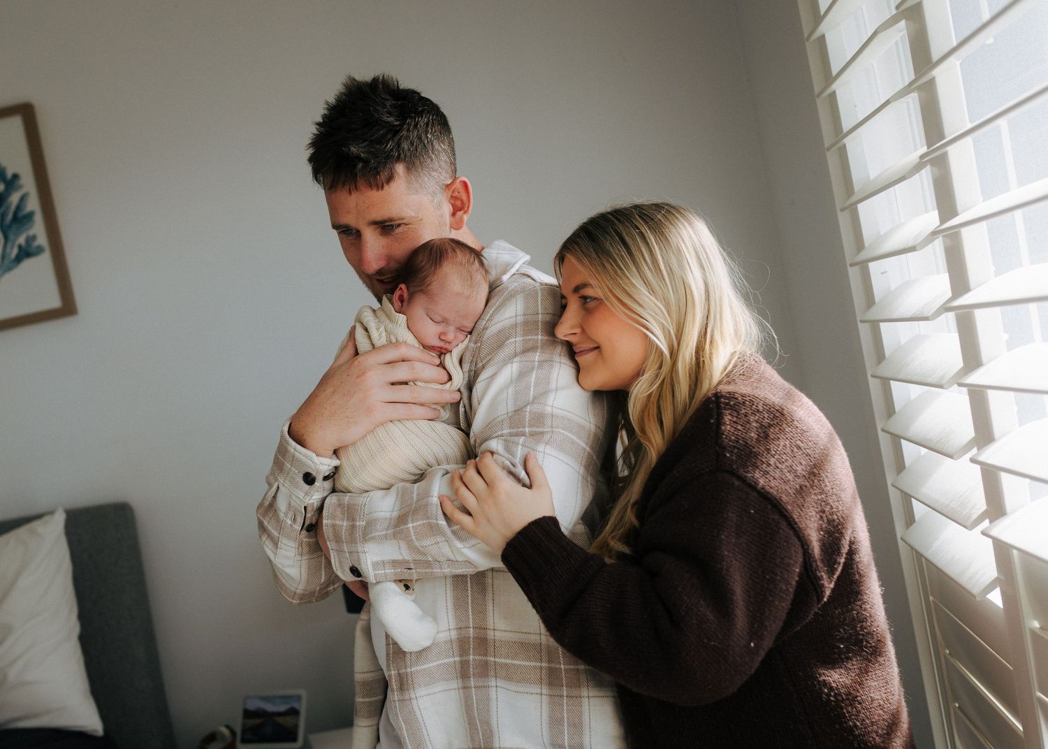 Smiling parents in plaid and brown sweaters holding newborn baby by window.