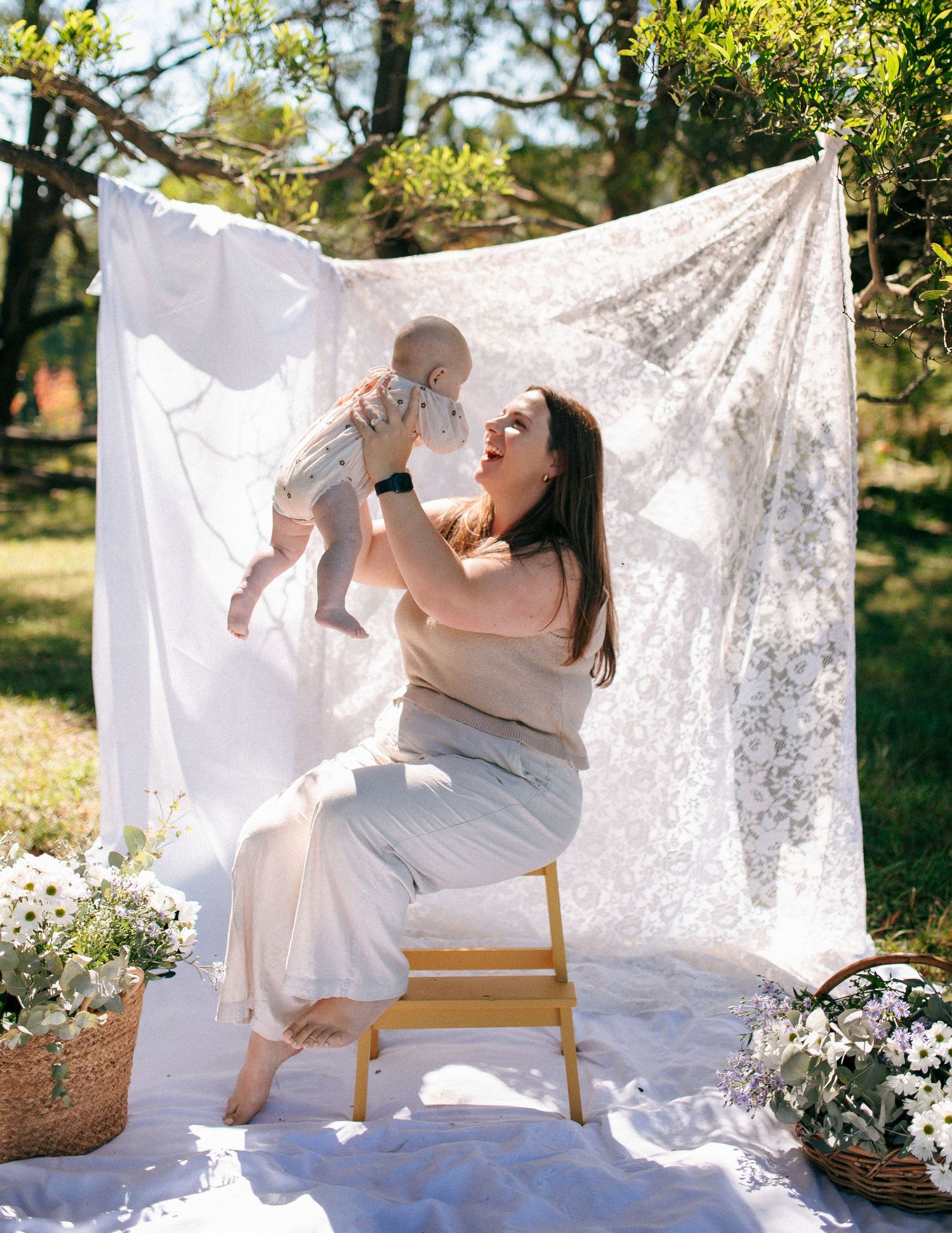Joyful mother lifting baby in air while seated outdoors with white flowers.