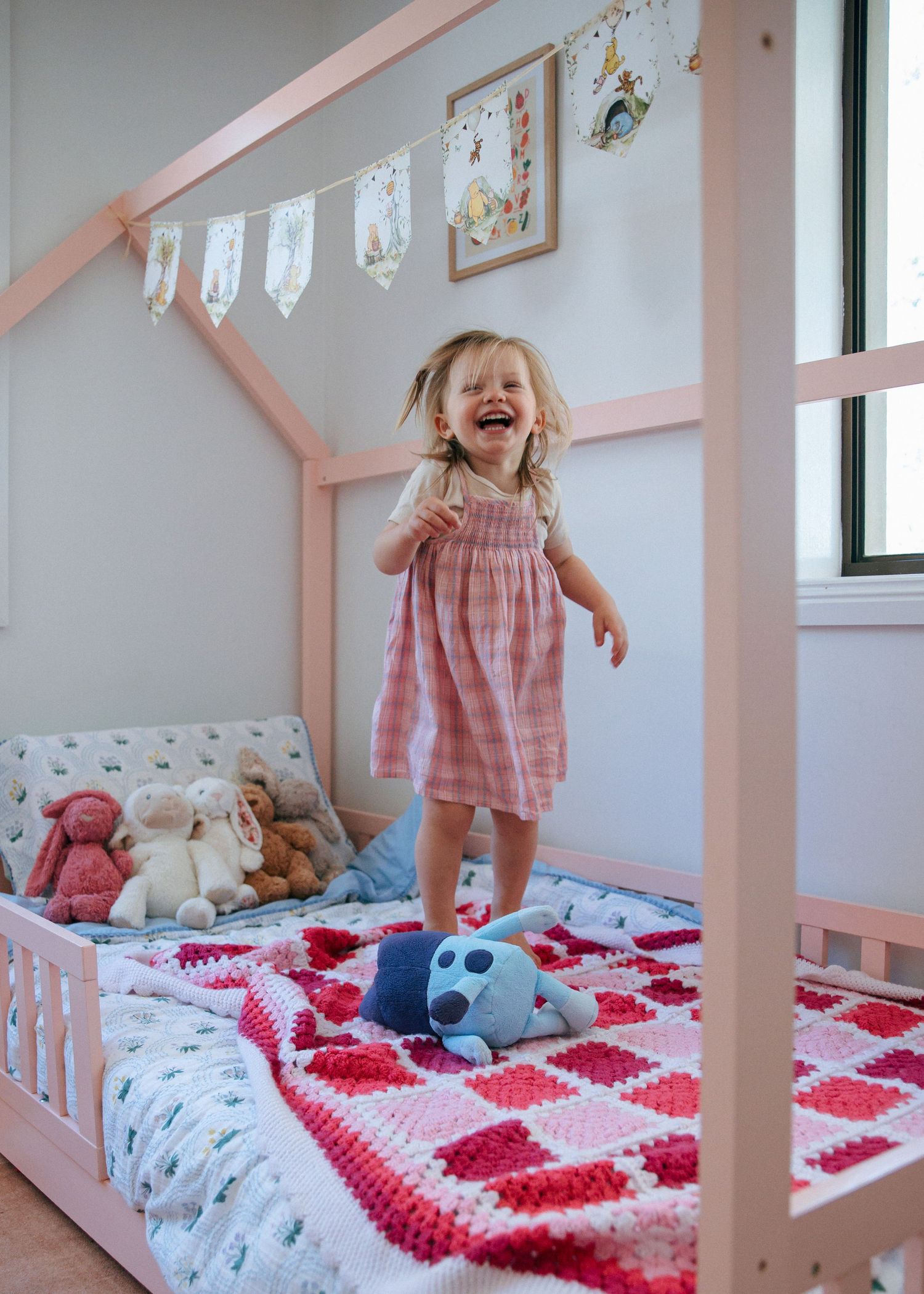 Laughing toddler girl jumping on a pink house bed frame with stuffed animals.