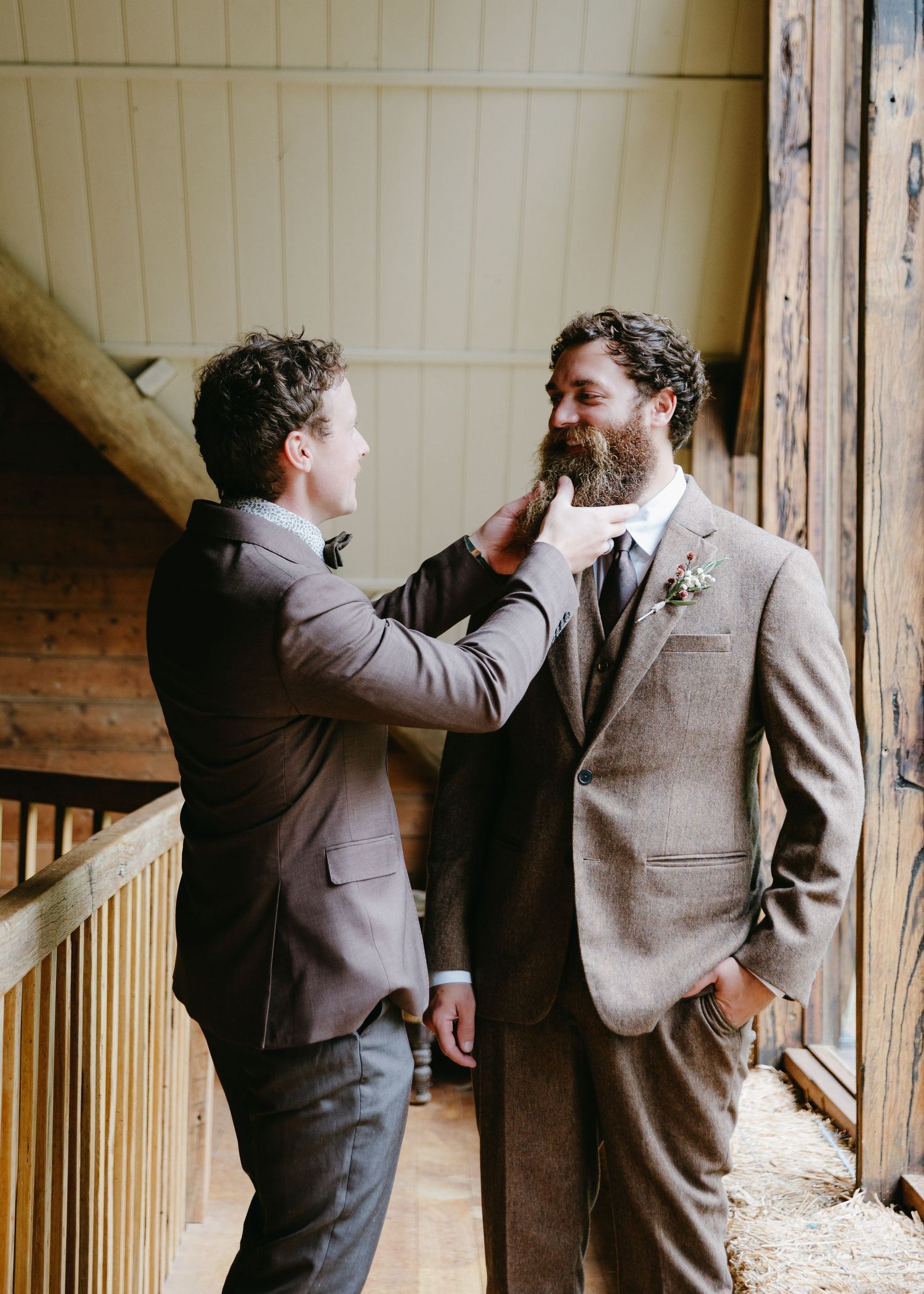 Groomsman adjusting bearded groom's boutonniere in a rustic barn before the wedding ceremony.