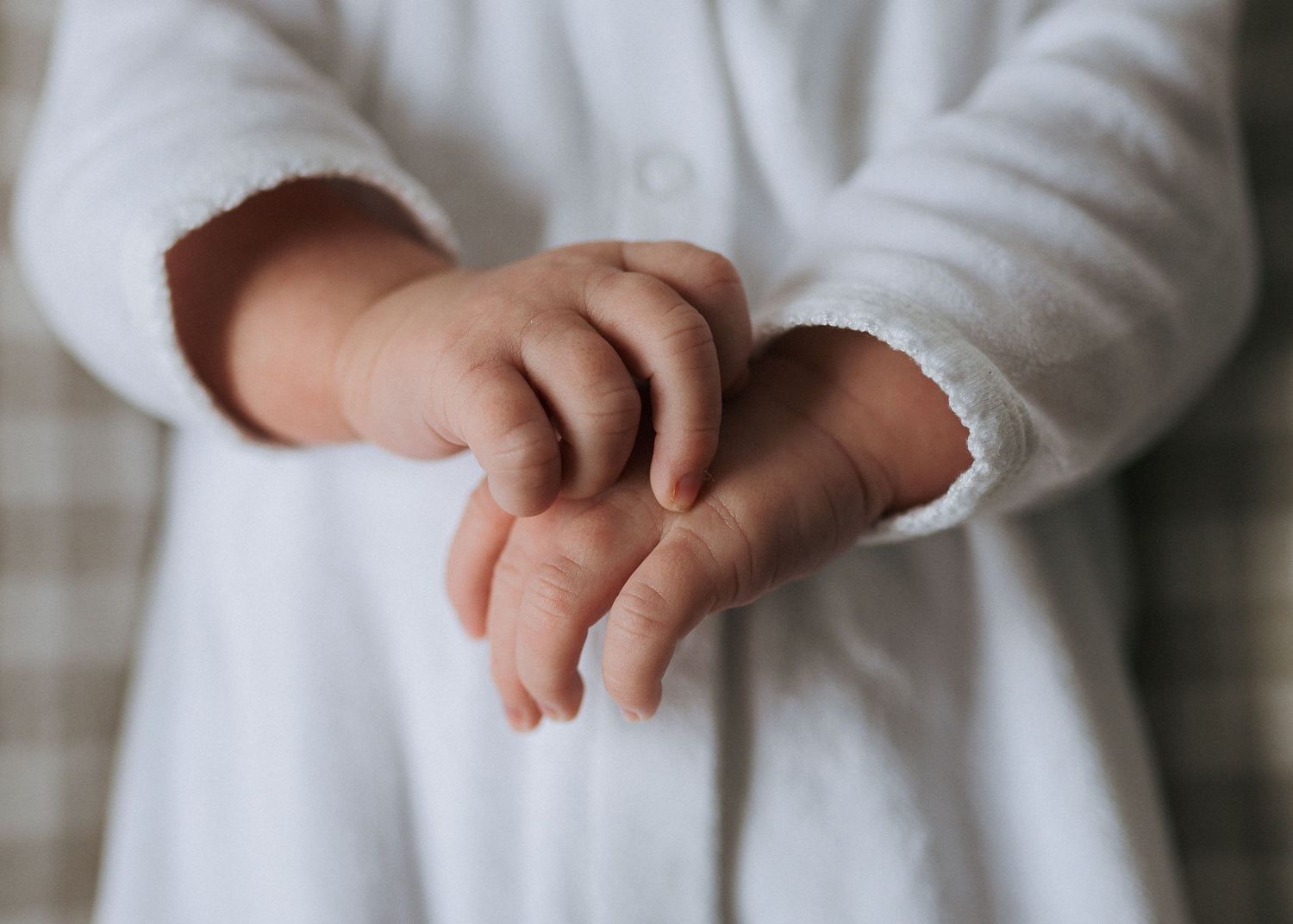 Tiny baby hands gently clasped together, dressed in a soft white cotton garment with a delicate scalloped cuff.