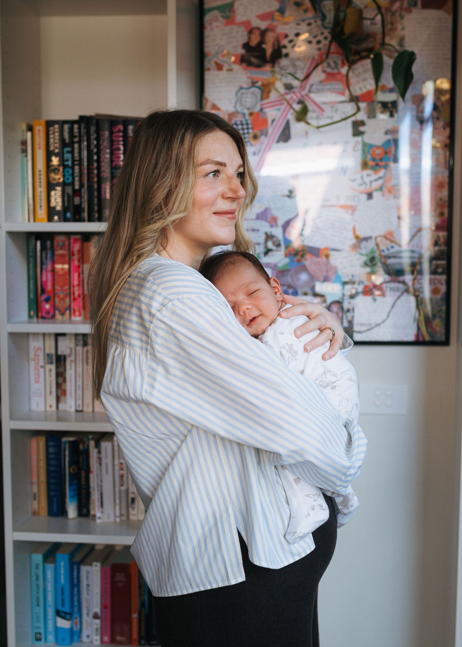 Woman with long blonde hair in a striped shirt cradles a sleeping newborn baby beside a full bookshelf and colorful framed art.