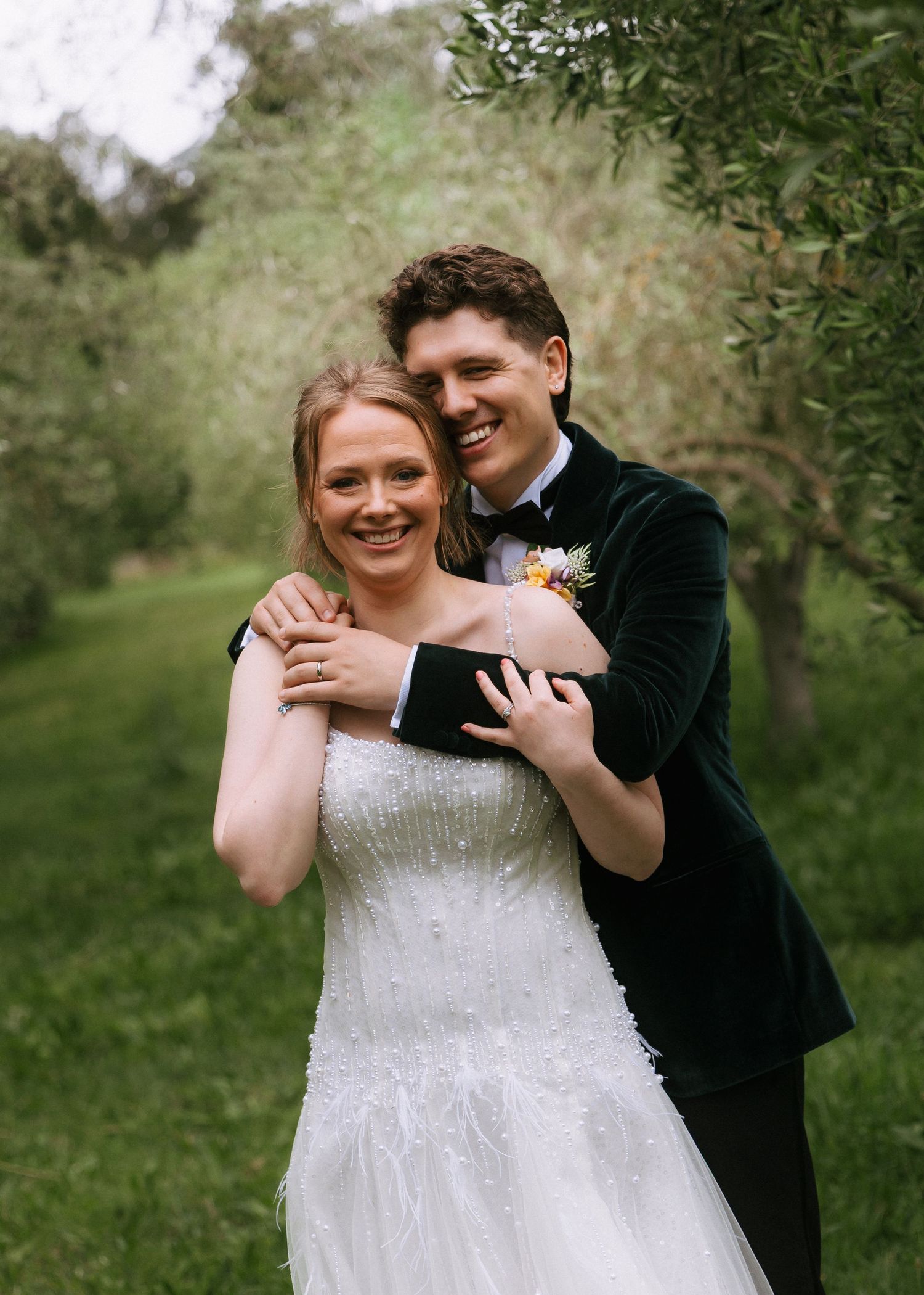 Smiling bride in a beaded white gown embraced from behind by a groom in a dark velvet jacket and bow tie among green olive trees.