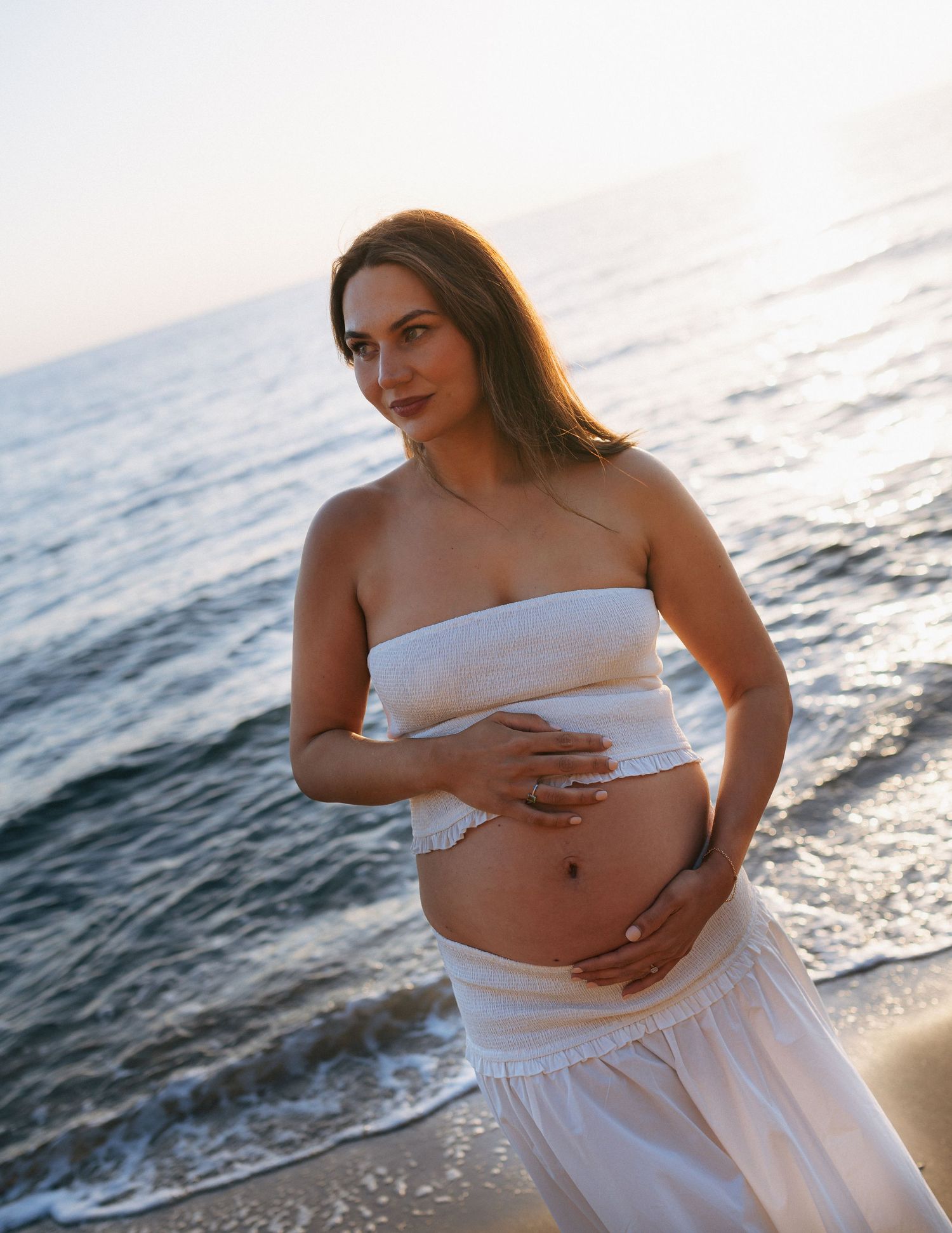 Pregnant woman in white tube top and skirt cradling her baby bump on a serene beach at sunset.