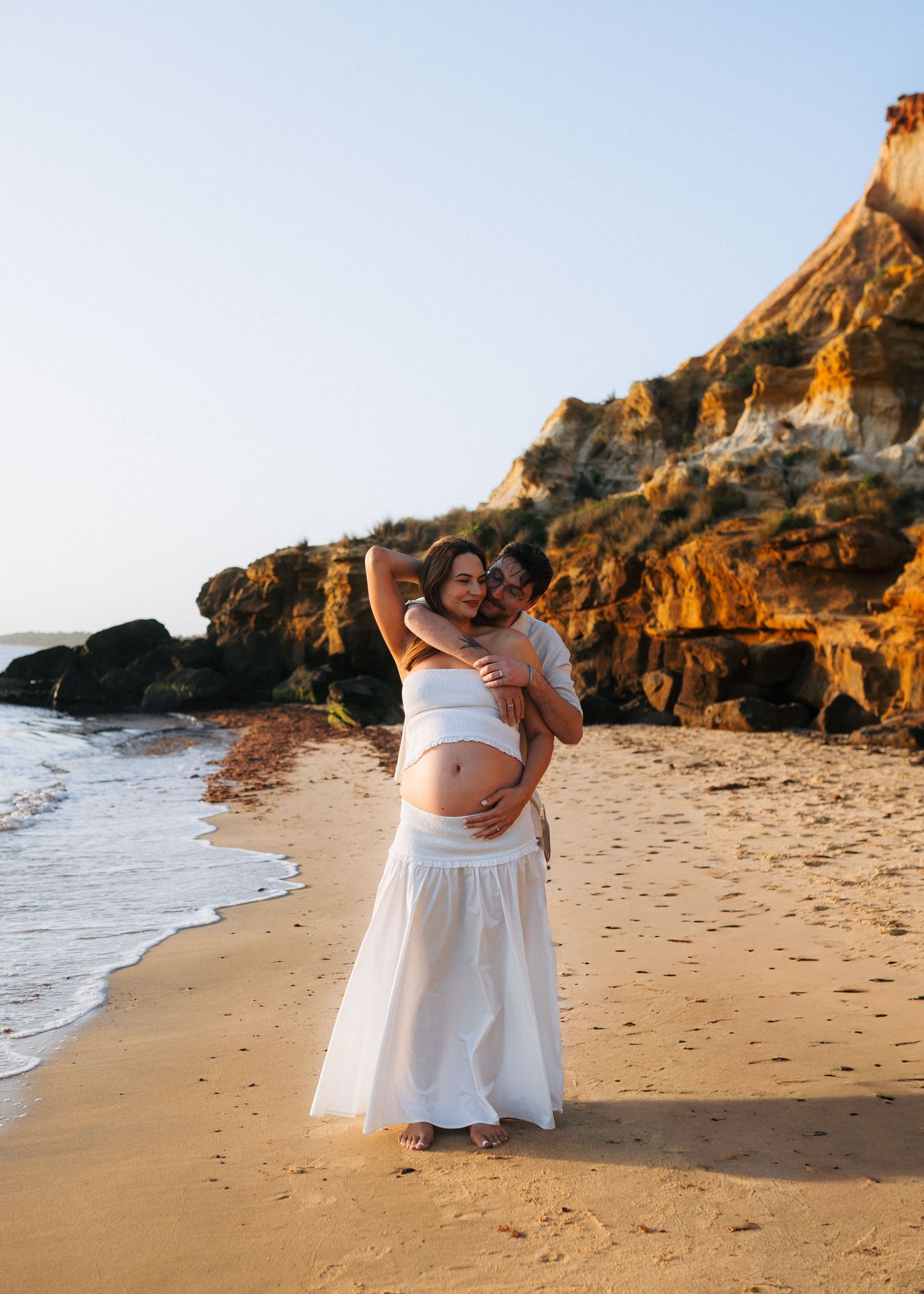 Expecting couple embraces on sandy beach at sunset, woman in white maternity dress showing baby bump.