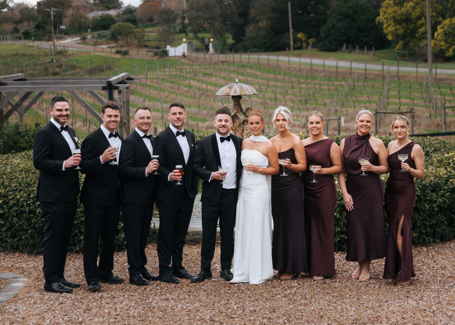 Wedding party in formal attire posing outdoors at a vineyard, groomsmen in tuxedos and bridesmaids in burgundy gowns.