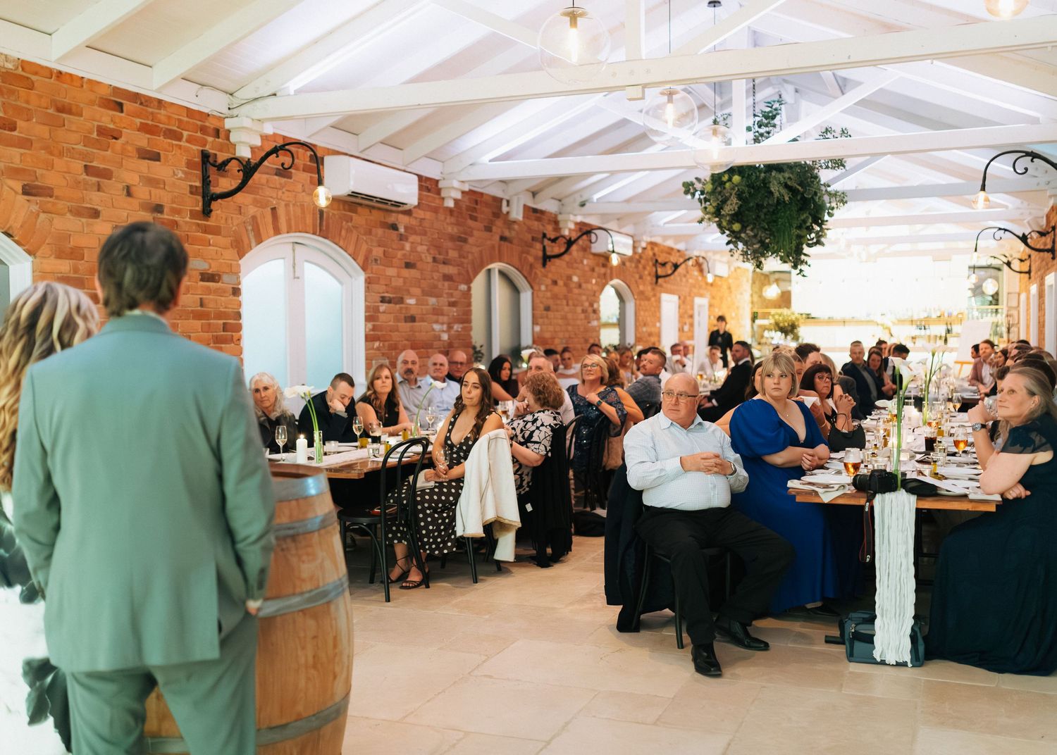 Wedding reception guests listen to speech in elegant brick venue with draped ceiling.