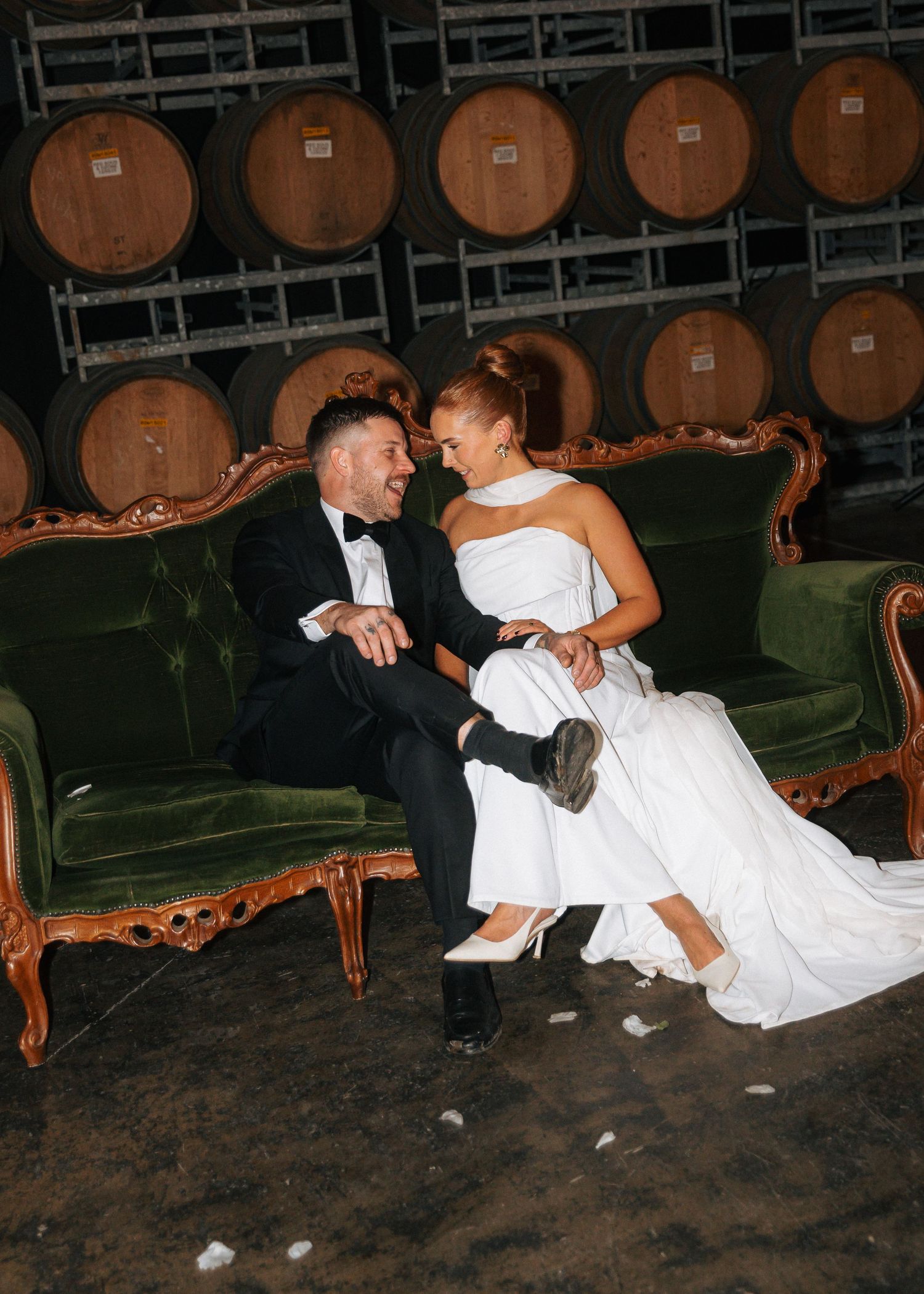 Bride and groom laughing together on a green velvet sofa in front of wine barrels at a winery wedding.