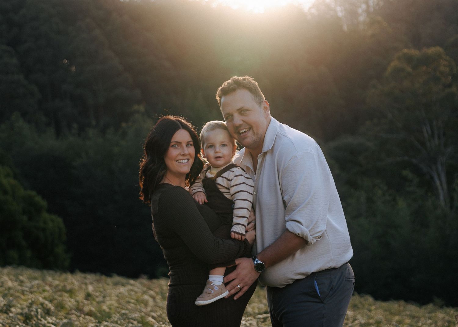 Happy family with pregnant mother and father holding laughing toddler in golden sunset field.
