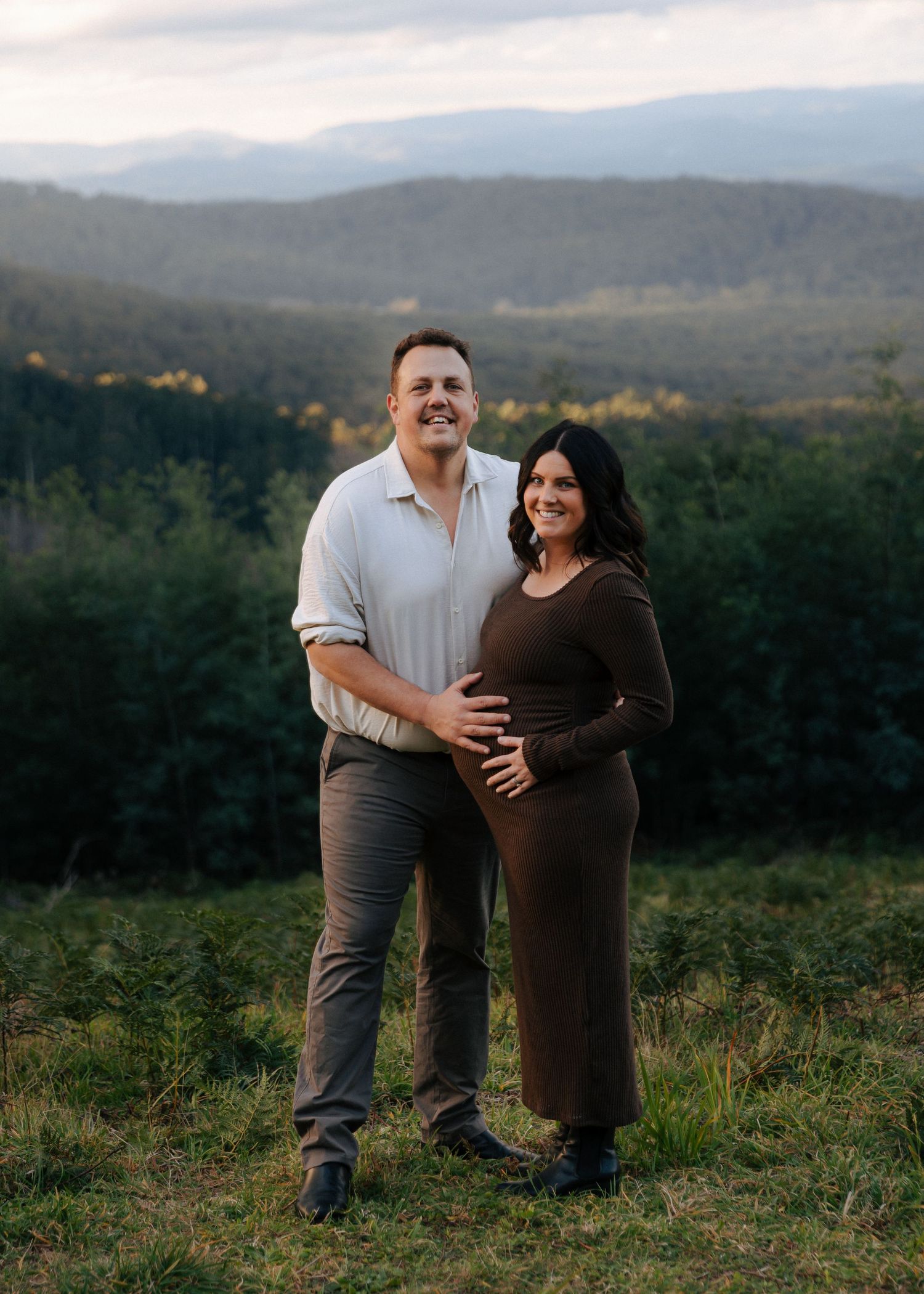 Smiling couple posing together in a green field with mountain views during maternity photoshoot.