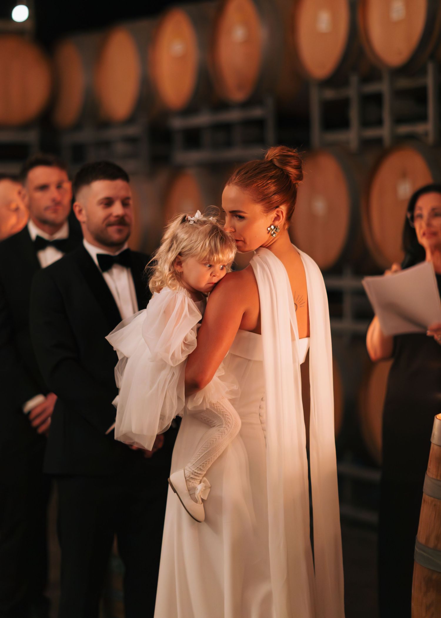 Bride in white halter gown tenderly holding young flower girl during wedding ceremony near wine barrels.