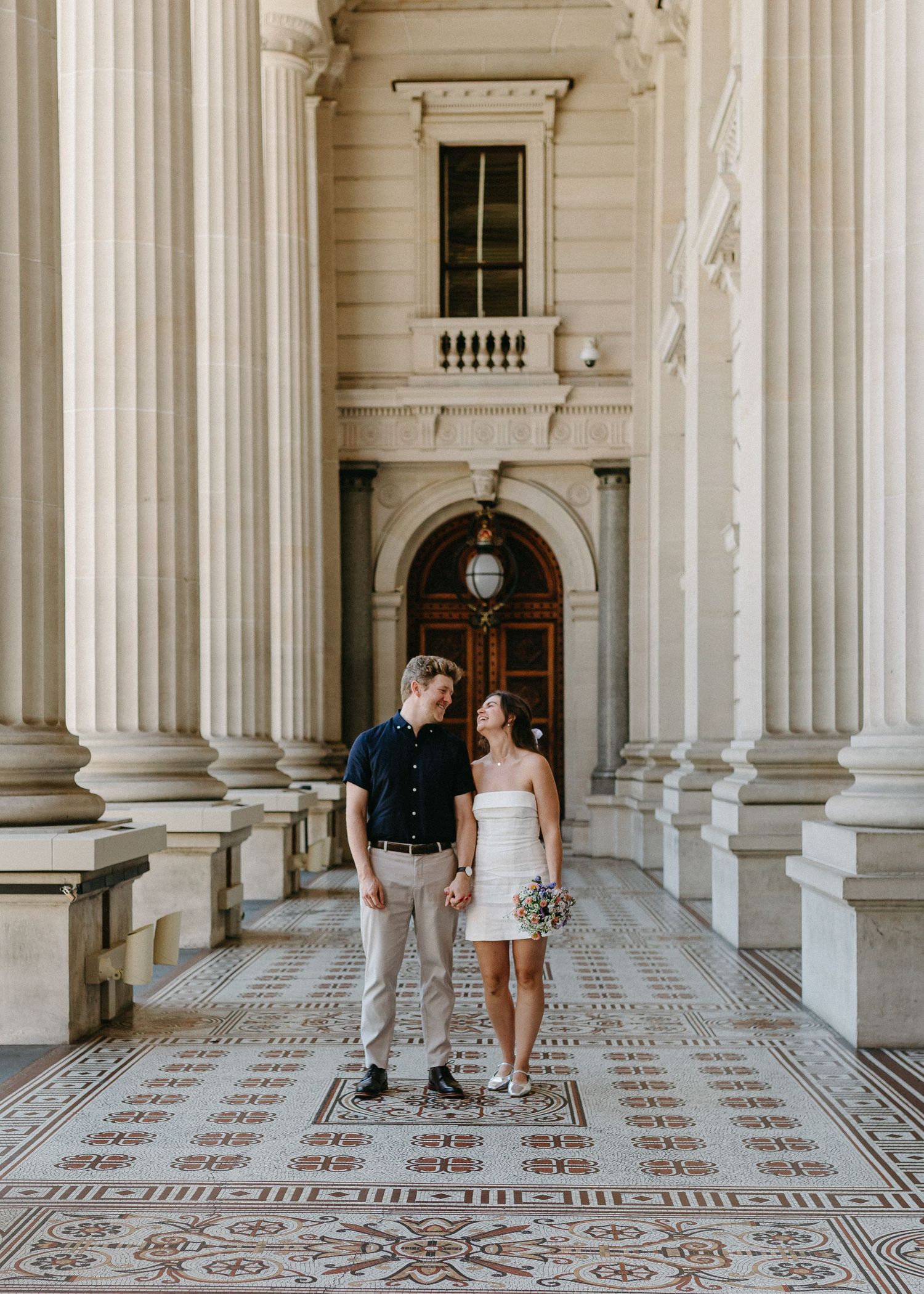 Bride and groom holding hands on ornate tiled floor surrounded by towering columns of a historic building.