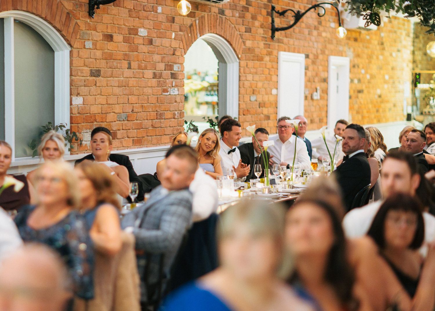 Guests seated at elegant wedding reception tables in a brick-walled venue with warm lighting and floral arrangements.