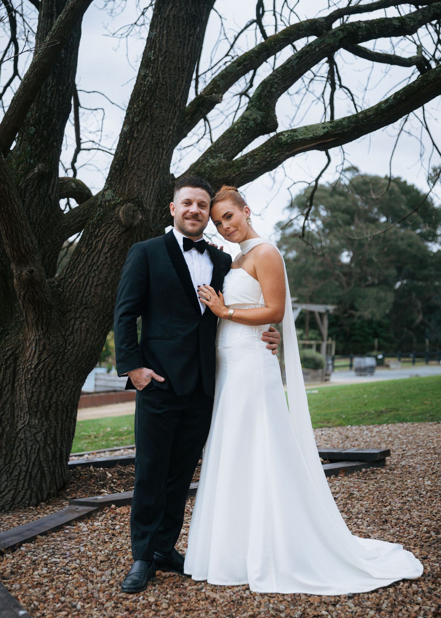 Bride and groom posing elegantly together under large tree in classic black tuxedo and white gown.