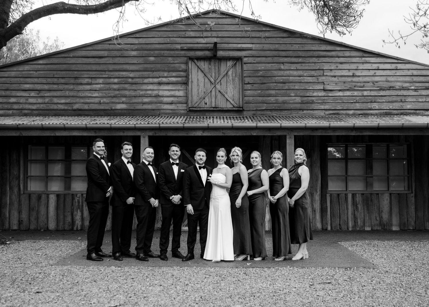 Wedding party poses in black and white photo outside rustic barn venue.