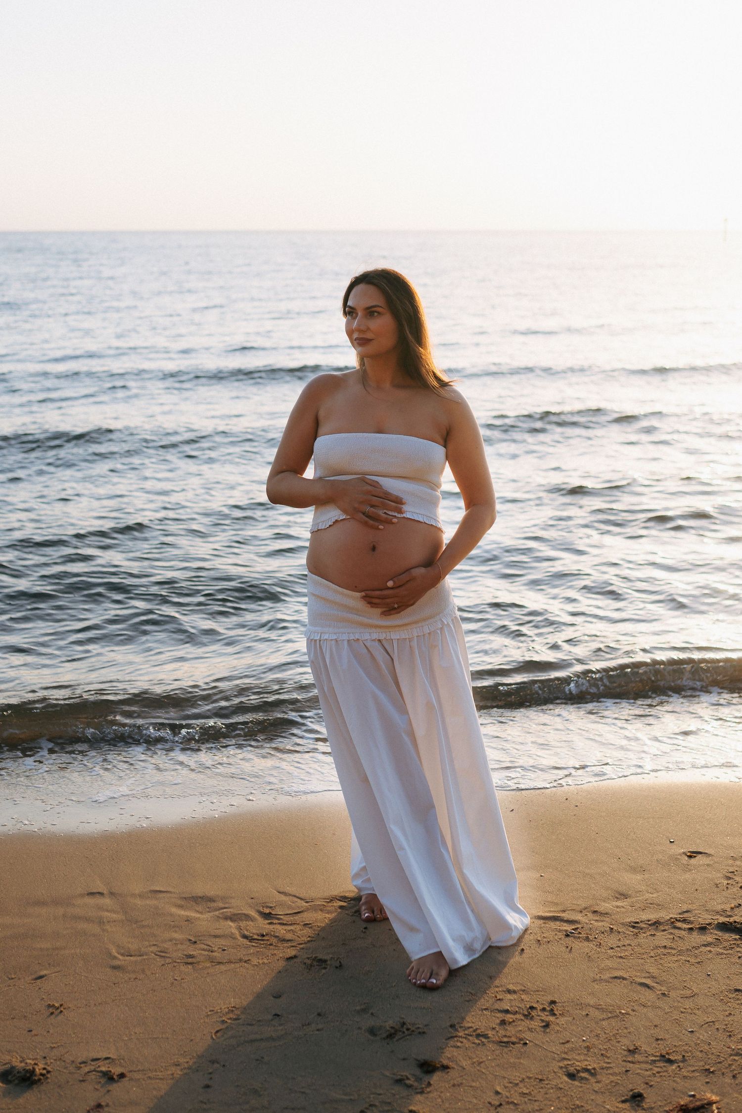 A pregnant woman in a white strapless top and skirt stands on a beach at sunset, cradling her belly.
