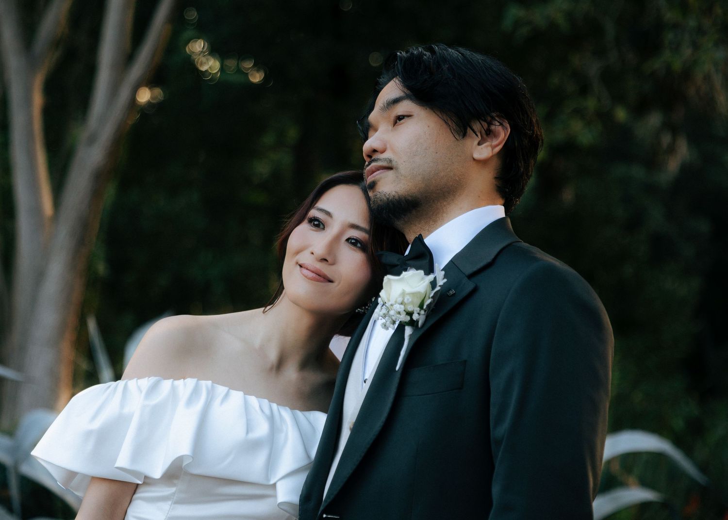 Close-up of bride and groom in elegant wedding attire looking upward together against a lush green backdrop.