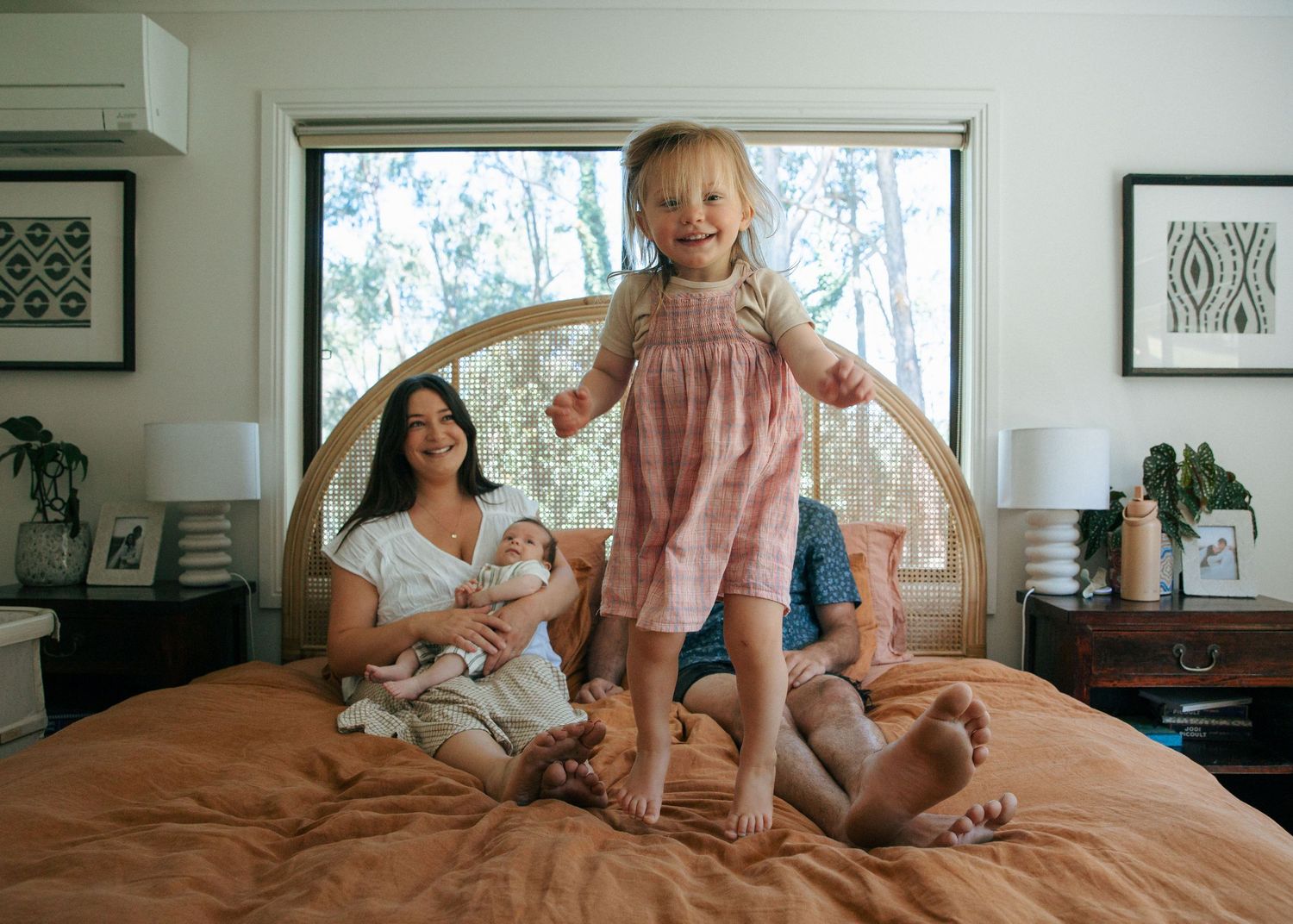 Mother holding newborn while toddler daughter stands smiling on orange bed.