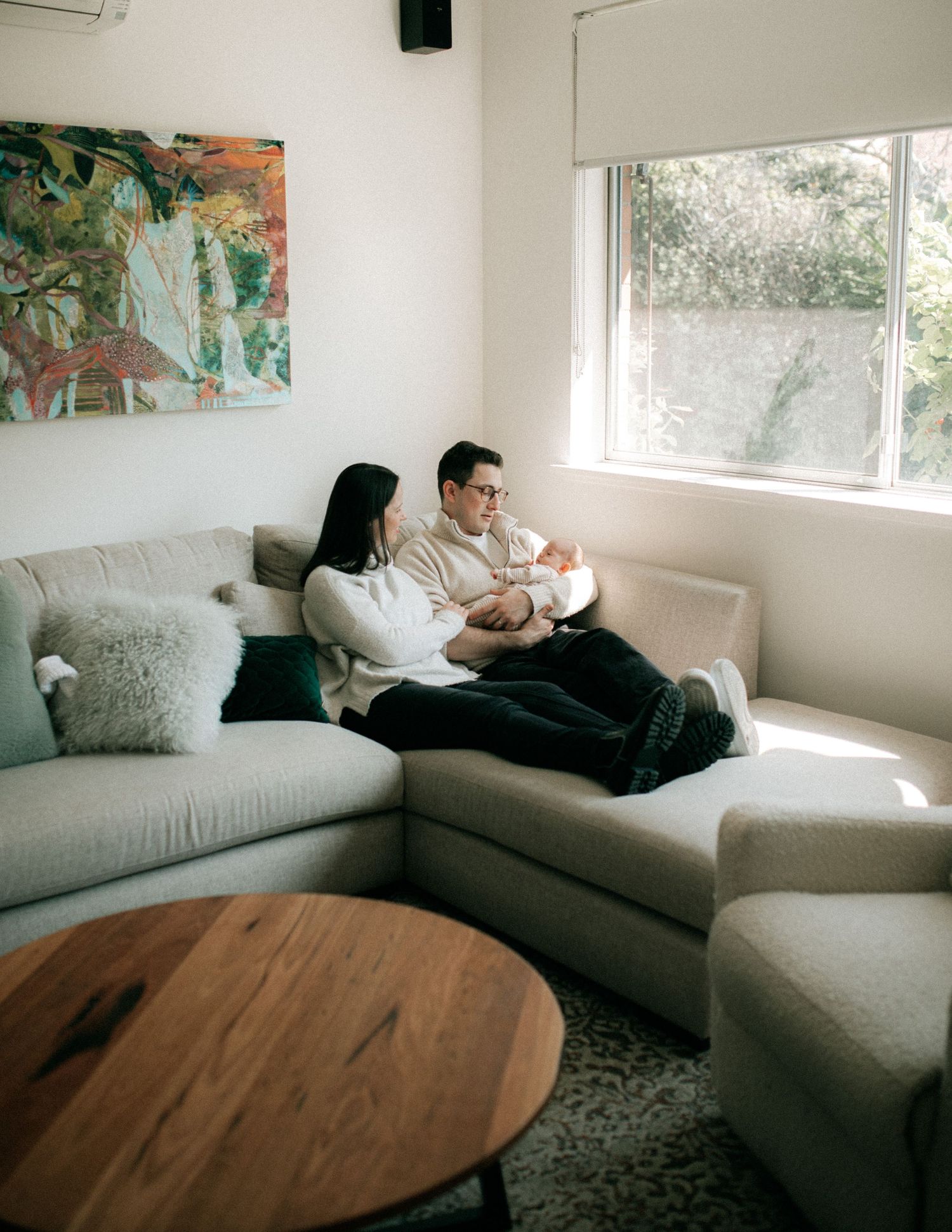 New parents sit together on a grey sectional sofa cradling their newborn baby by a bright window.