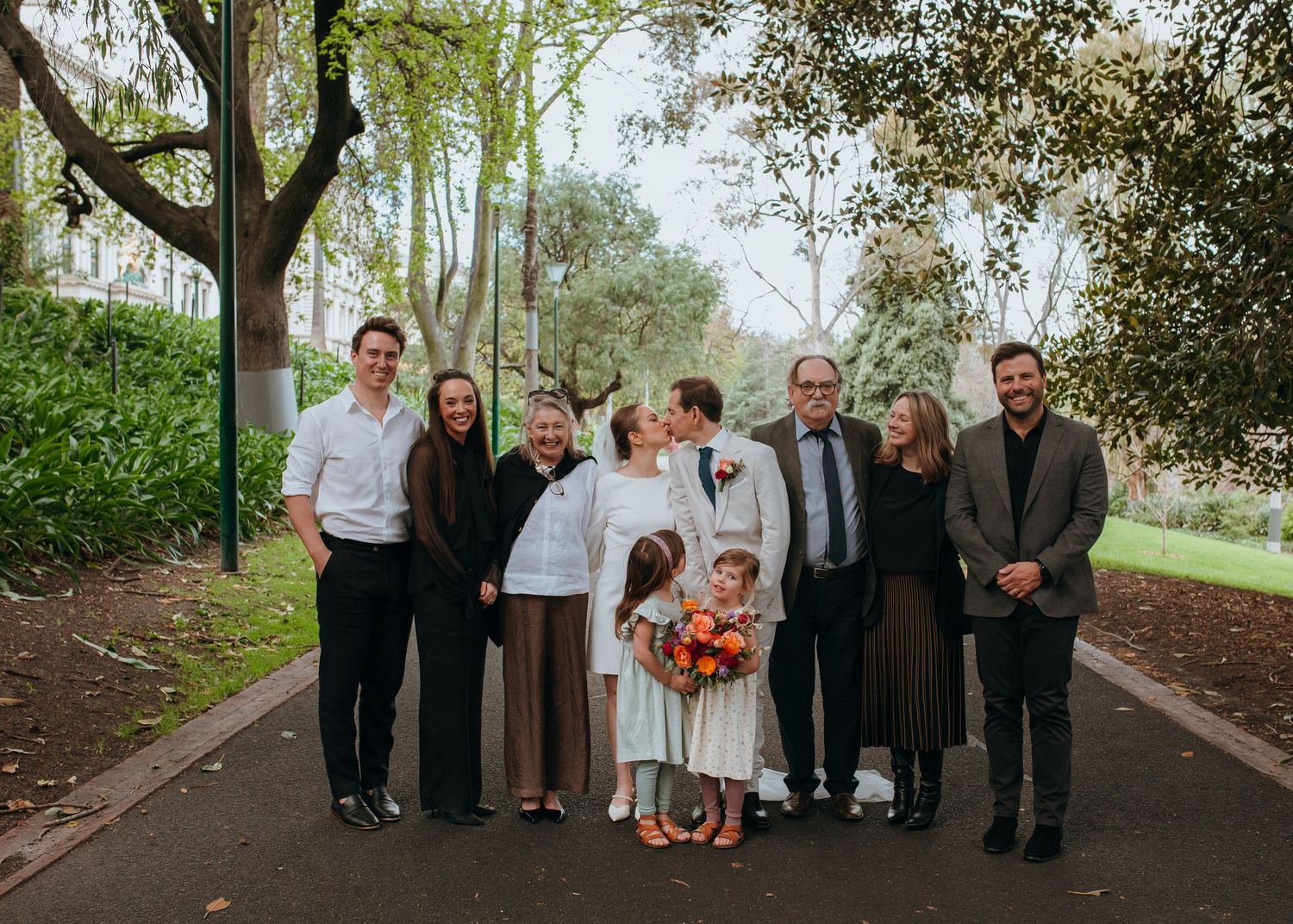 Wedding party group photo on a tree-lined park path with bride in white dress holding orange bouquet.