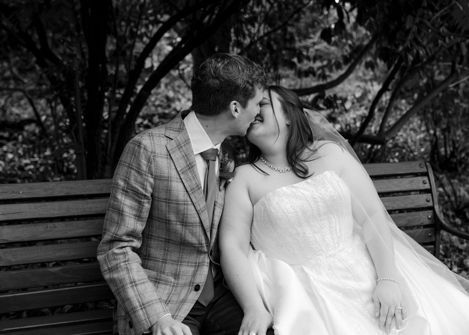 Black and white photo of bride and groom sharing a kiss on a bench in a garden.