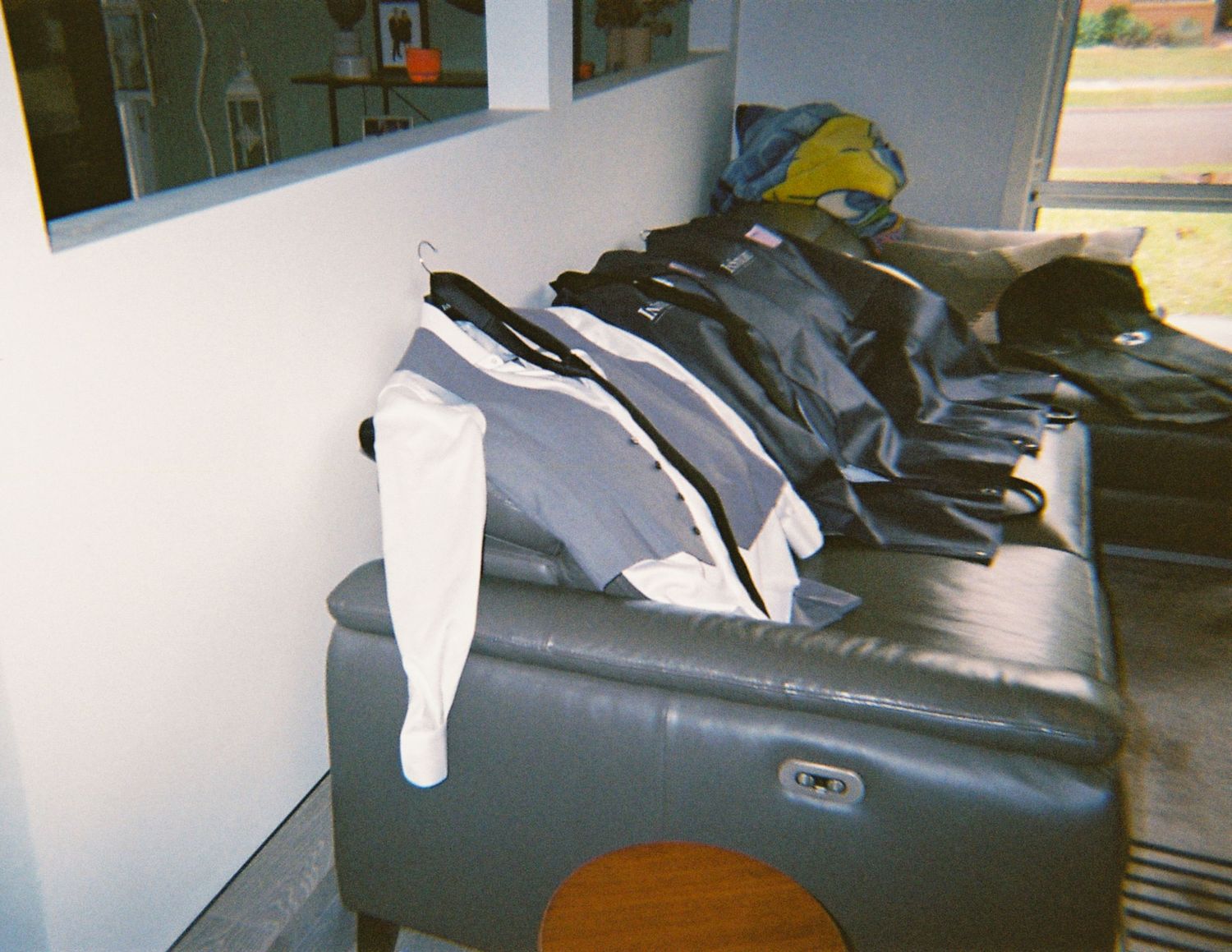 Groomsmen suits and jackets draped over a dark leather couch in a preparation room.