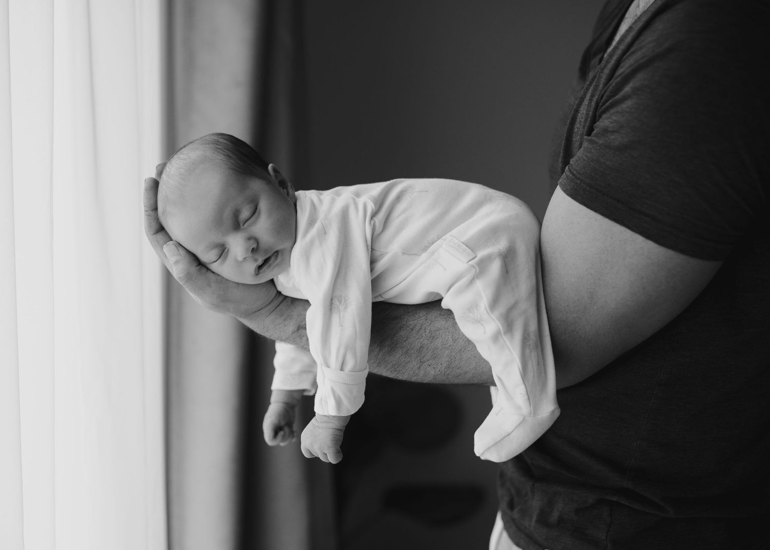 Black and white photo of newborn baby resting on father's arm near window.