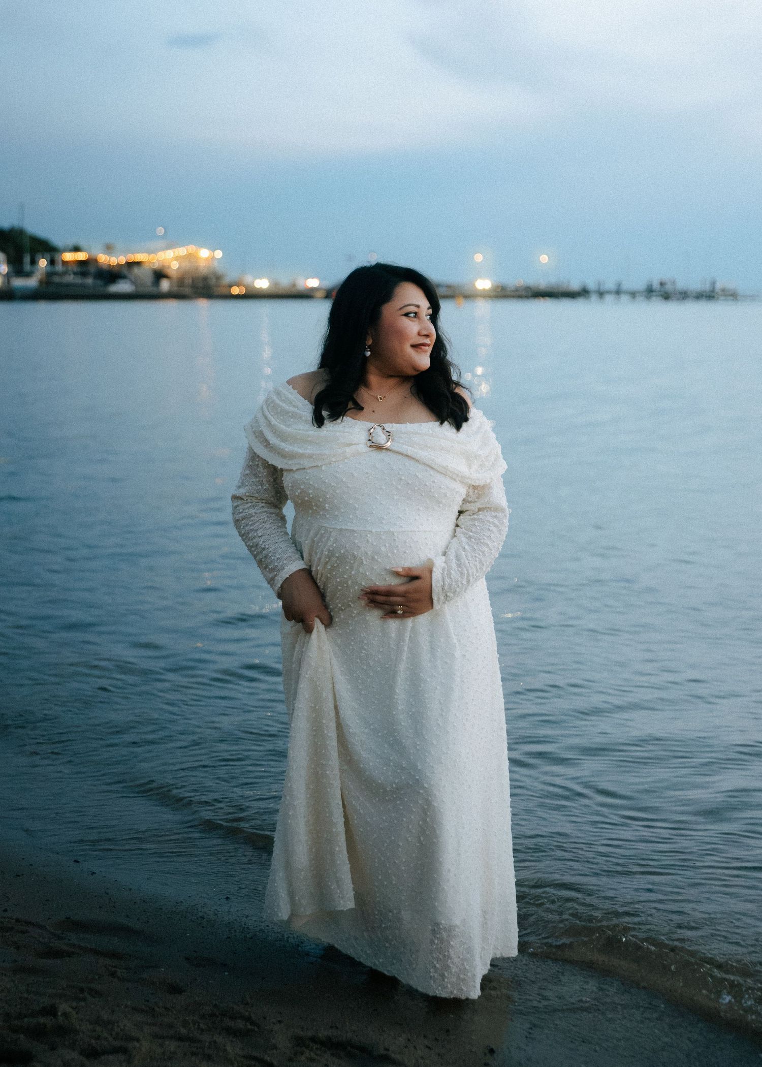 Pregnant woman in white off-shoulder lace dress cradling belly near waterfront at dusk.