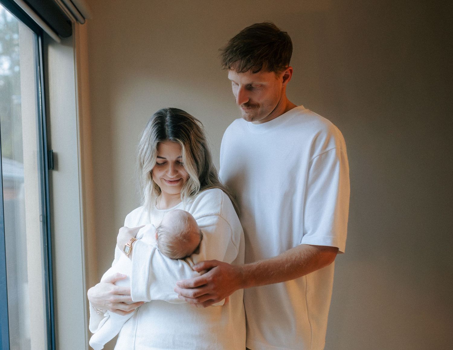 New parents tenderly looking down at their newborn baby held by mother near window.