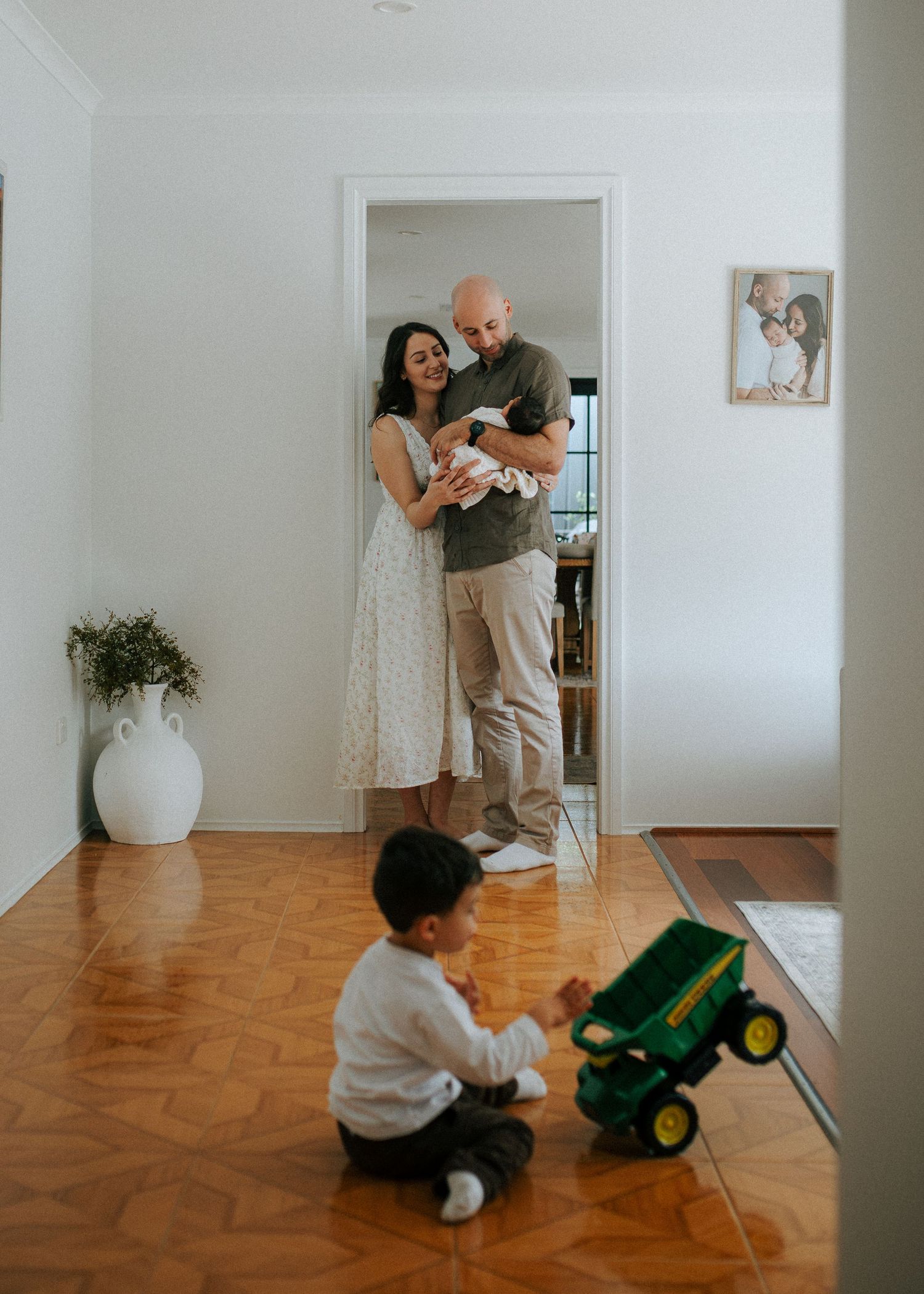 Parents in doorway with newborn while toddler plays with green toy truck on wood floor.