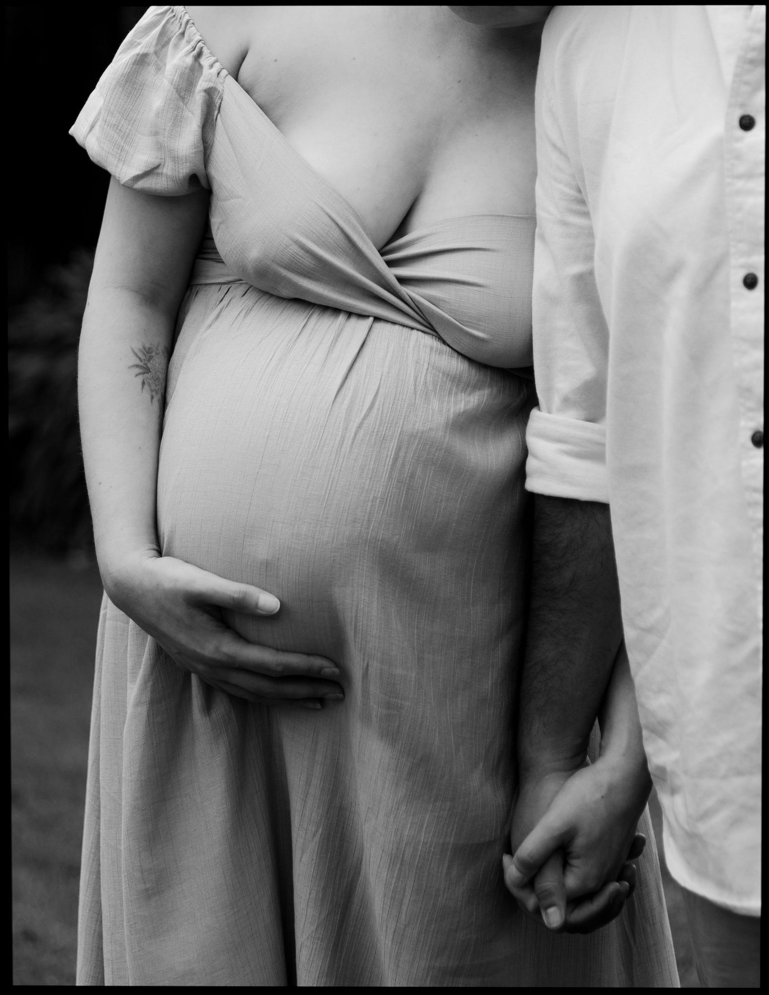 Black and white photo of pregnant couple holding hands, woman's baby bump visible in flowy dress.