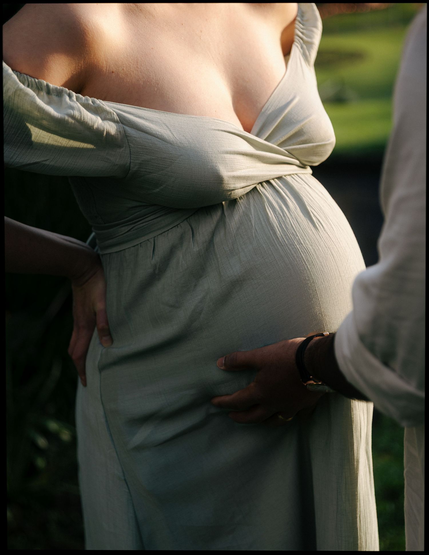 Close-up maternity photo of pregnant belly in sage green dress with partner's hand resting on her back.