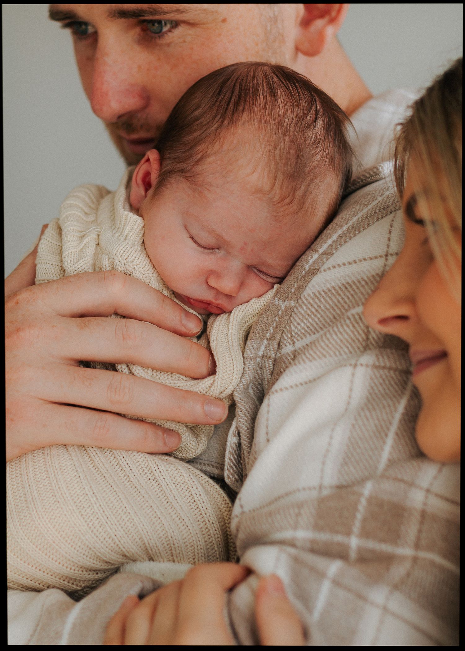 Parents tenderly embracing sleeping newborn baby wrapped in soft cream knit blanket.