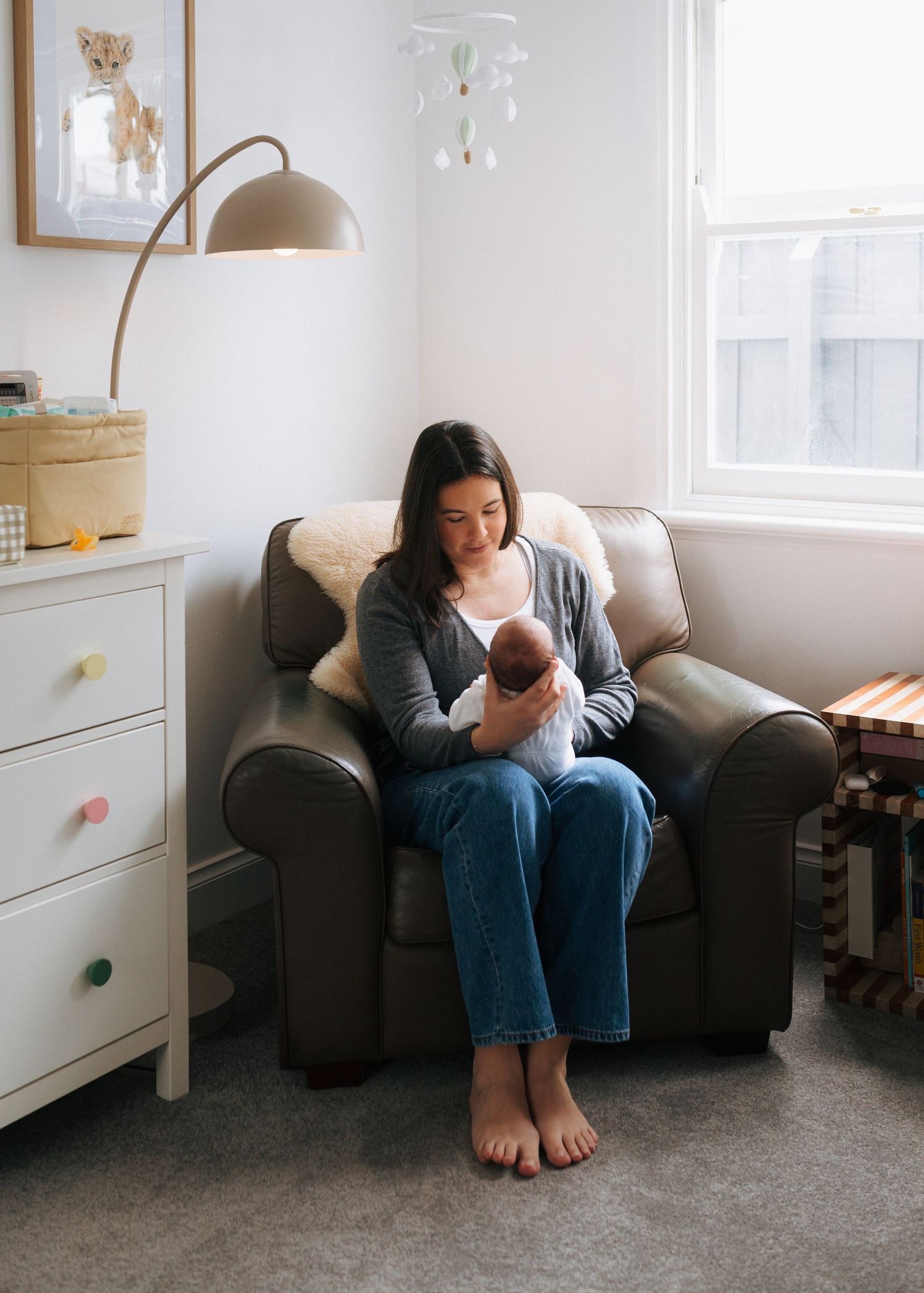 Barefoot mother in jeans sits in a leather armchair holding her sleeping newborn baby in a bright, cozy nursery corner.