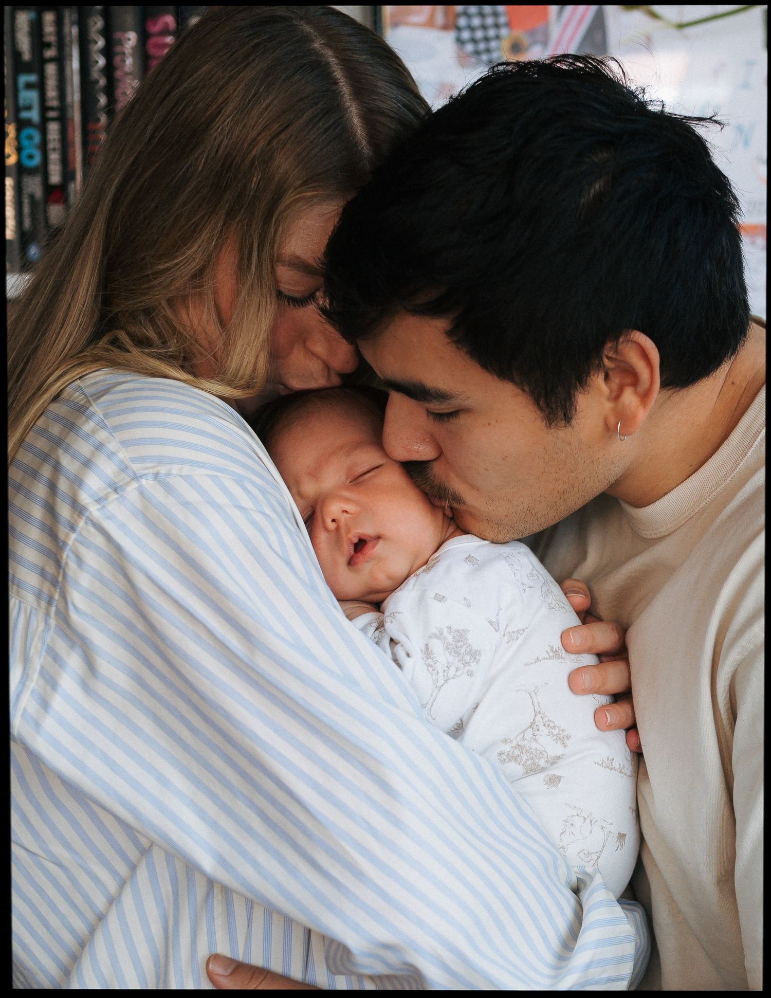 Parents tenderly kiss their sleeping newborn baby's cheeks while cradling the infant in a cozy, intimate family moment.