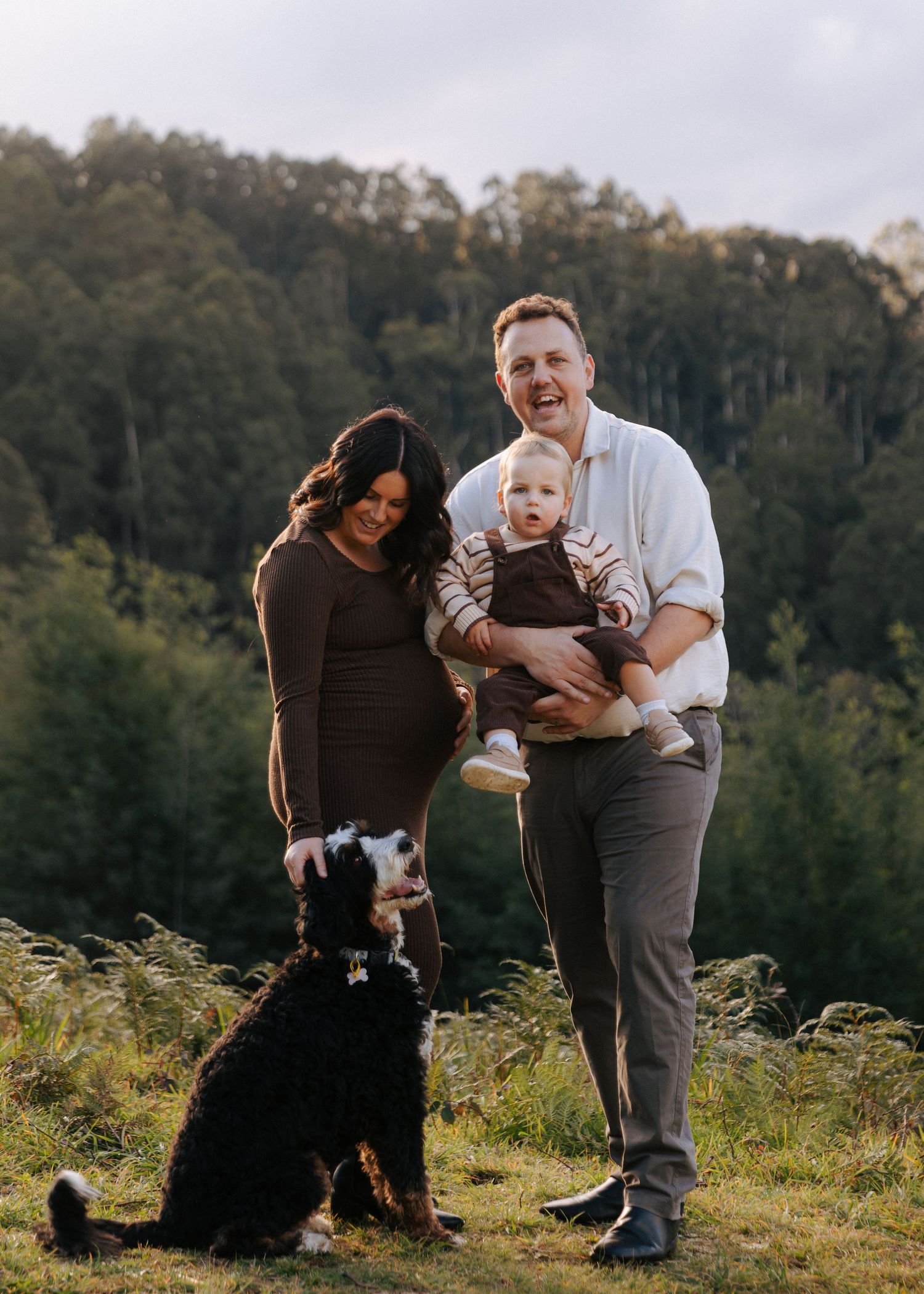 Laughing family with pregnant mom, dad holding toddler, and black dog on lush green hill.
