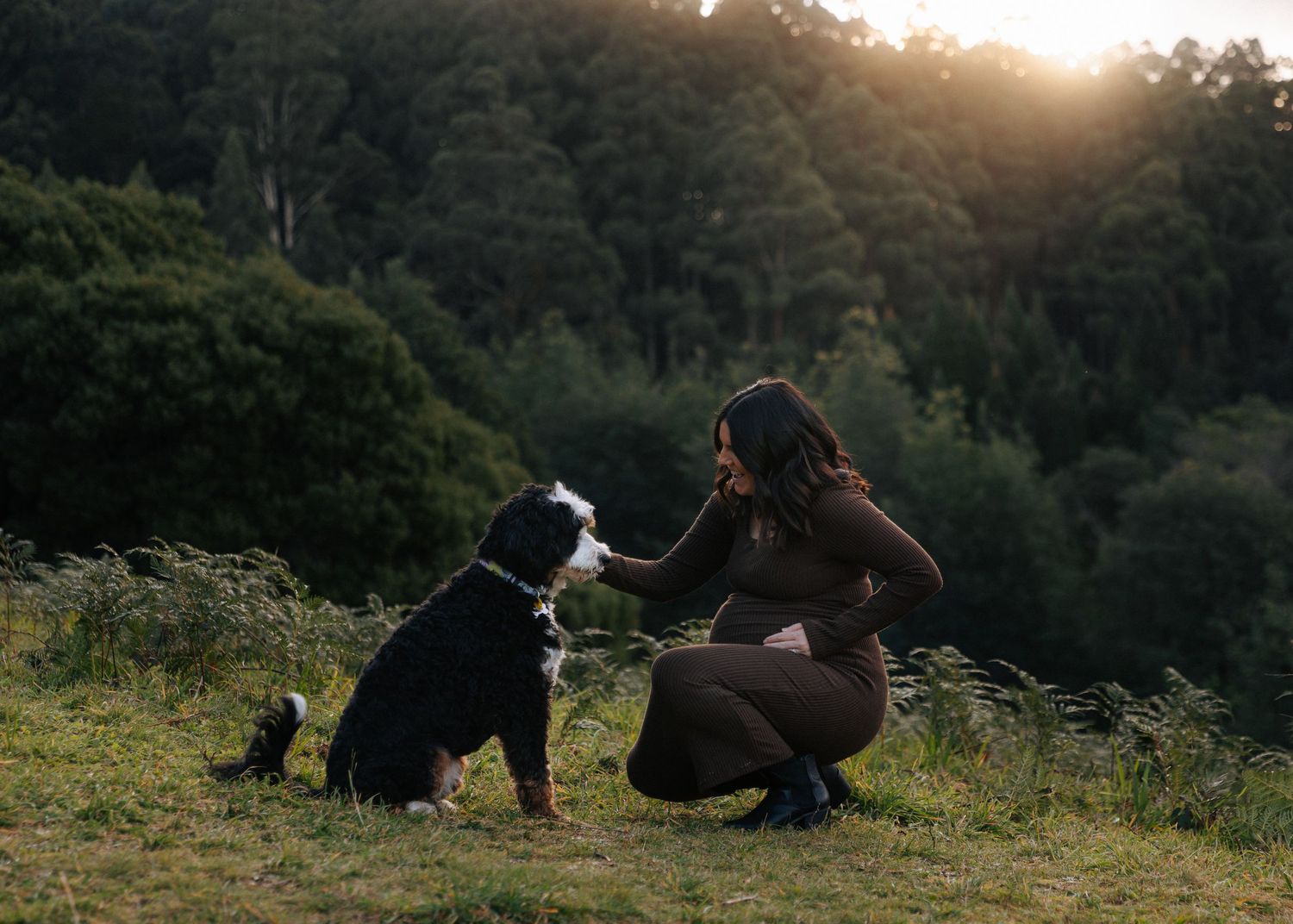 Pregnant woman in brown outfit kneeling to interact with a black and white dog at sunset.